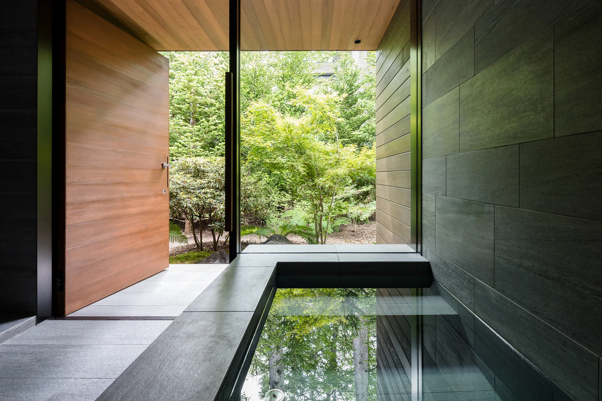 A modern indoor spa featuring a grey stone soaking tub next to a large open wooden door that looks out onto a green leafy garden.