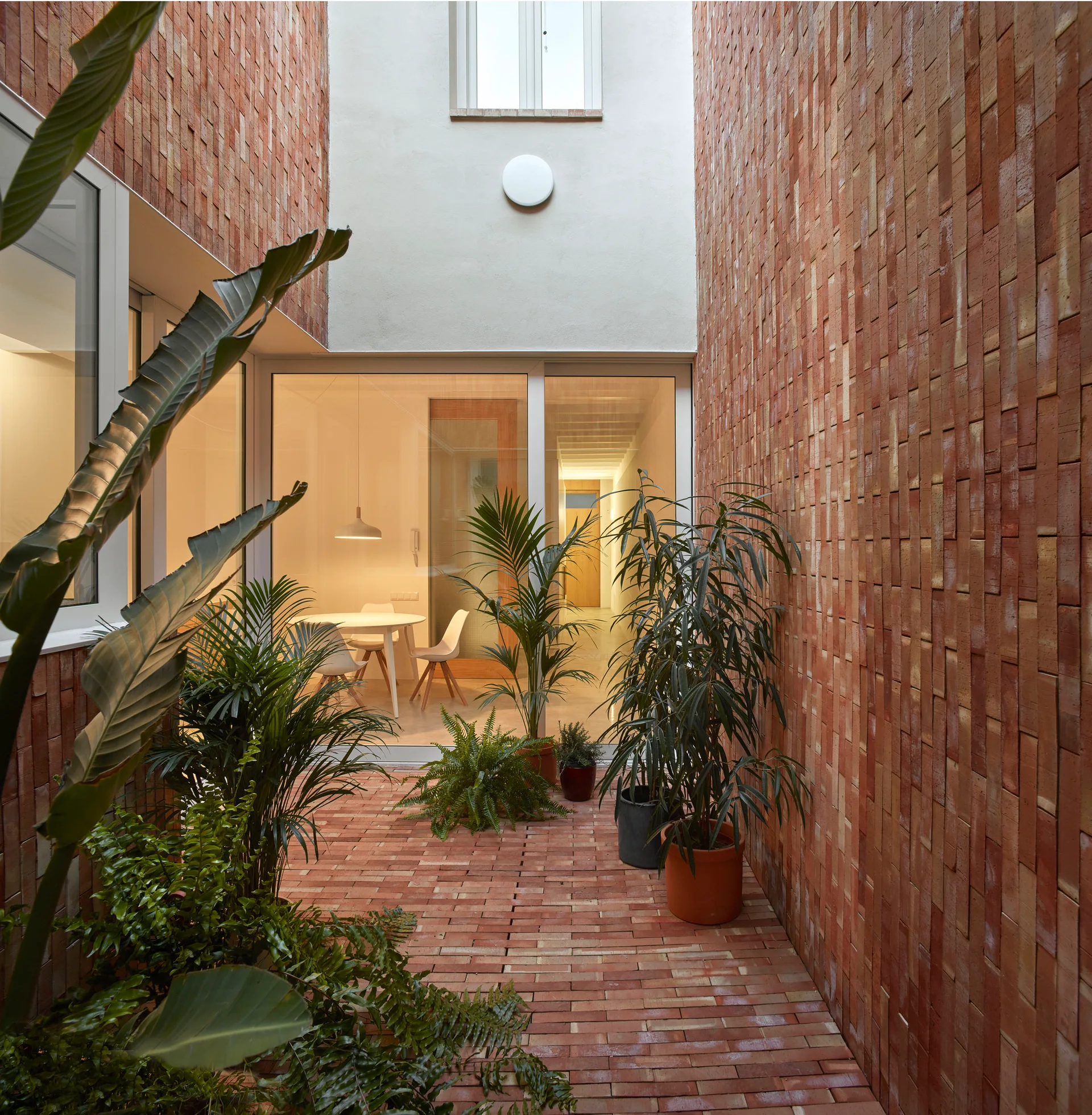A narrow brick courtyard with terracotta walls and potted plants looks into a modern dining room through large sliding glass doors.