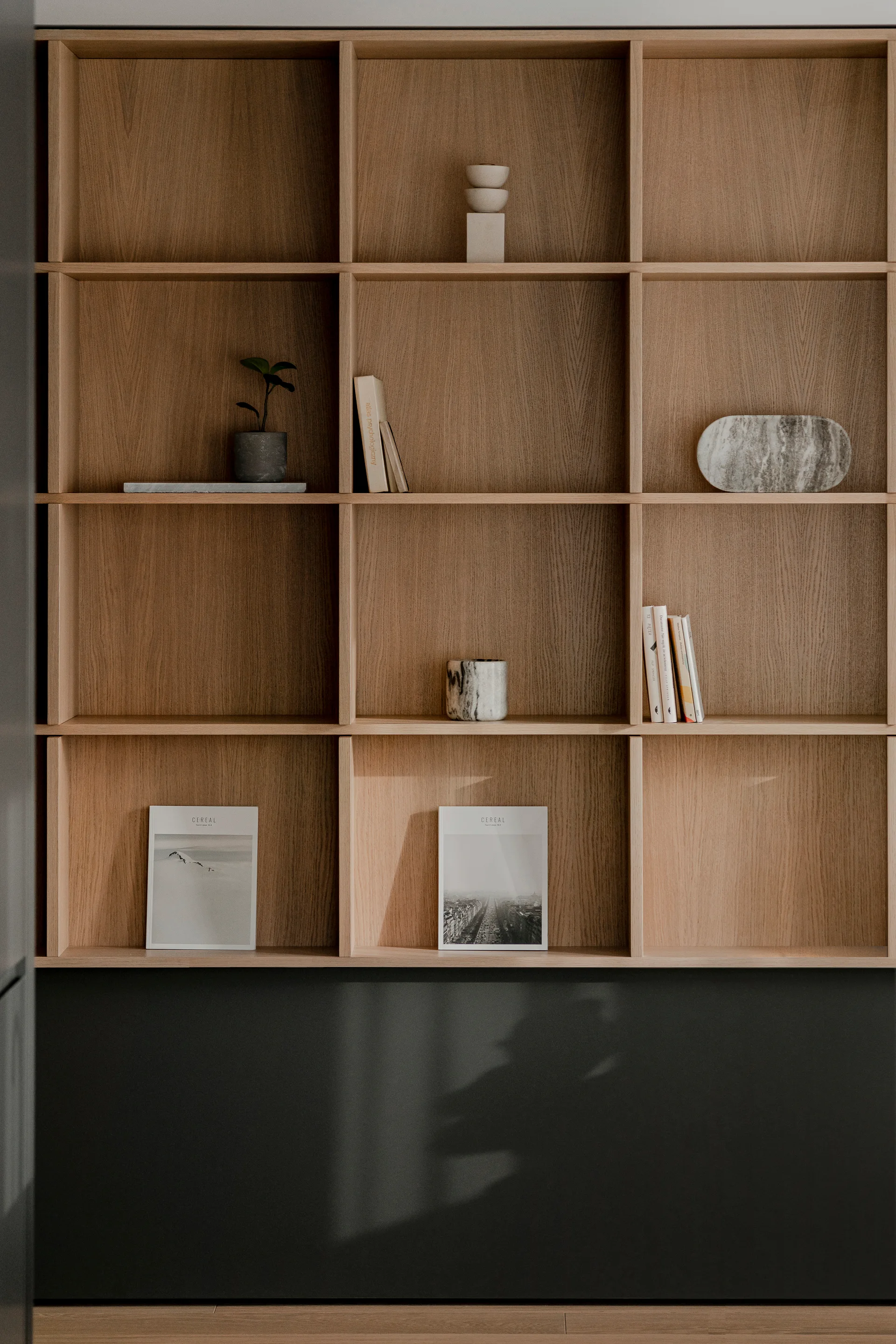 A natural wood grid shelving unit styled with books, ceramic objects, and a small plant above a dark grey base cabinet.