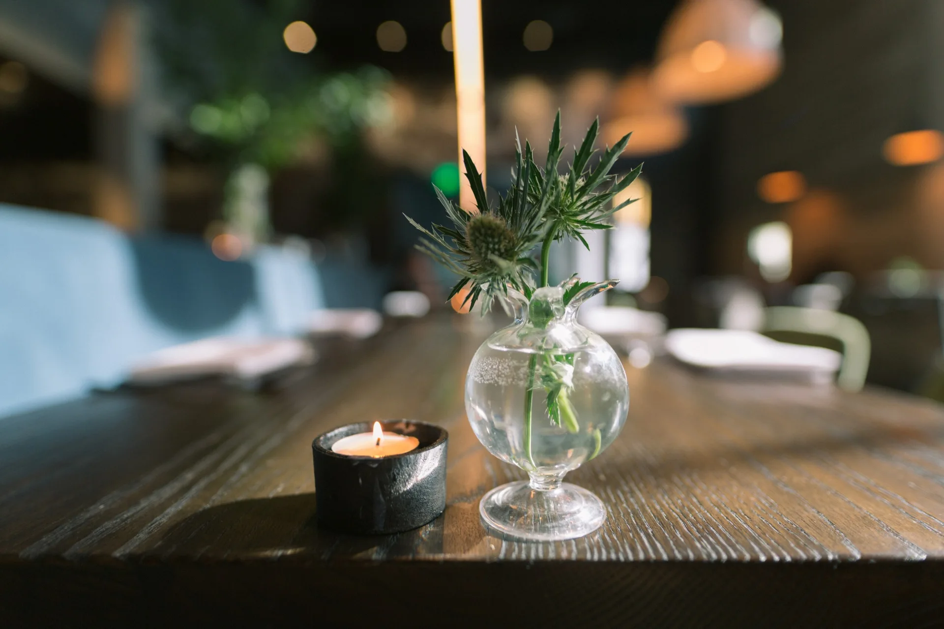 A close-up of a dark wood dining table featuring a small clear glass vase with spiky green foliage next to a lit black votive candle.