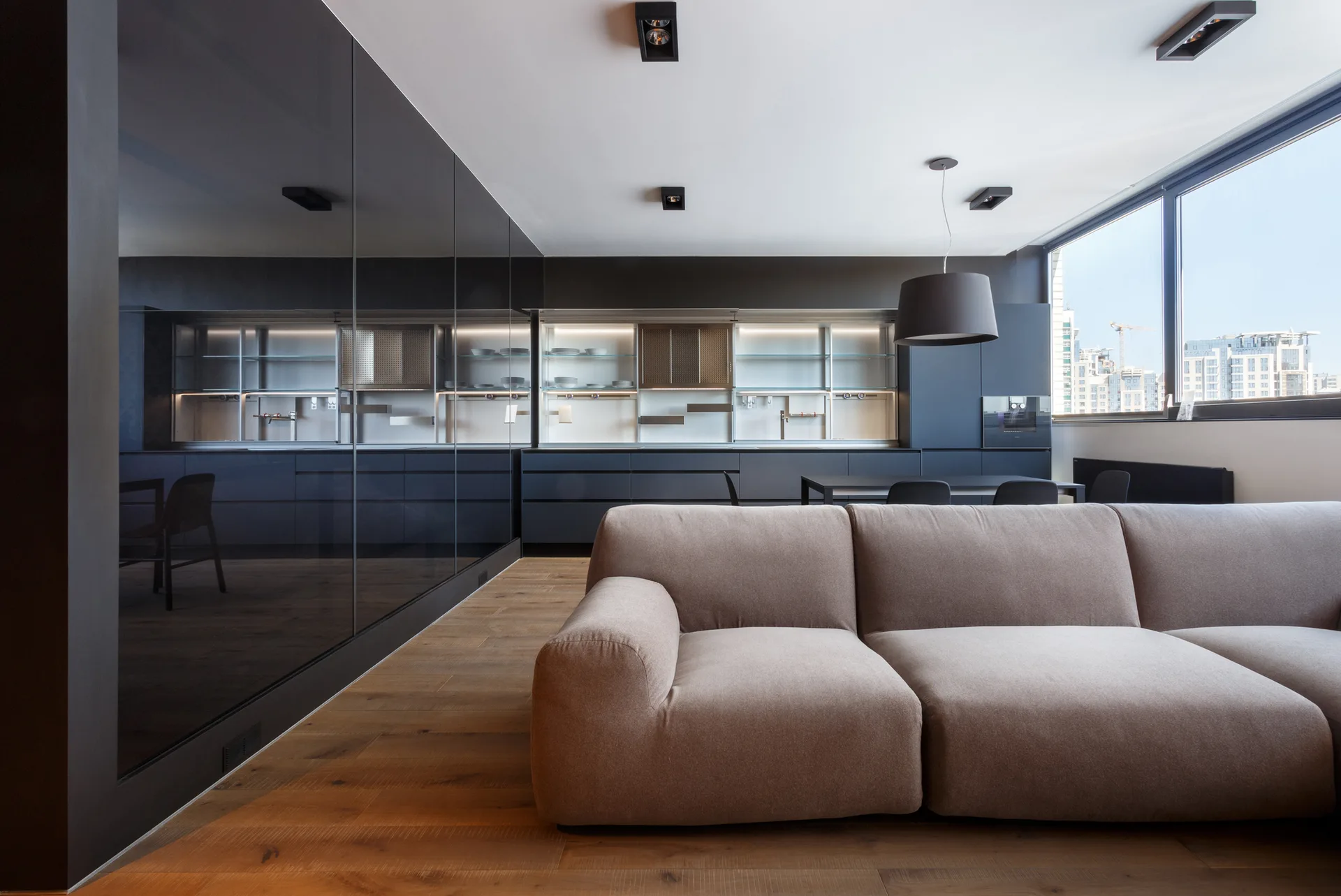 A contemporary open-plan room features a low tan sectional sofa on light wood floors beside a large reflective black glass accent wall.
