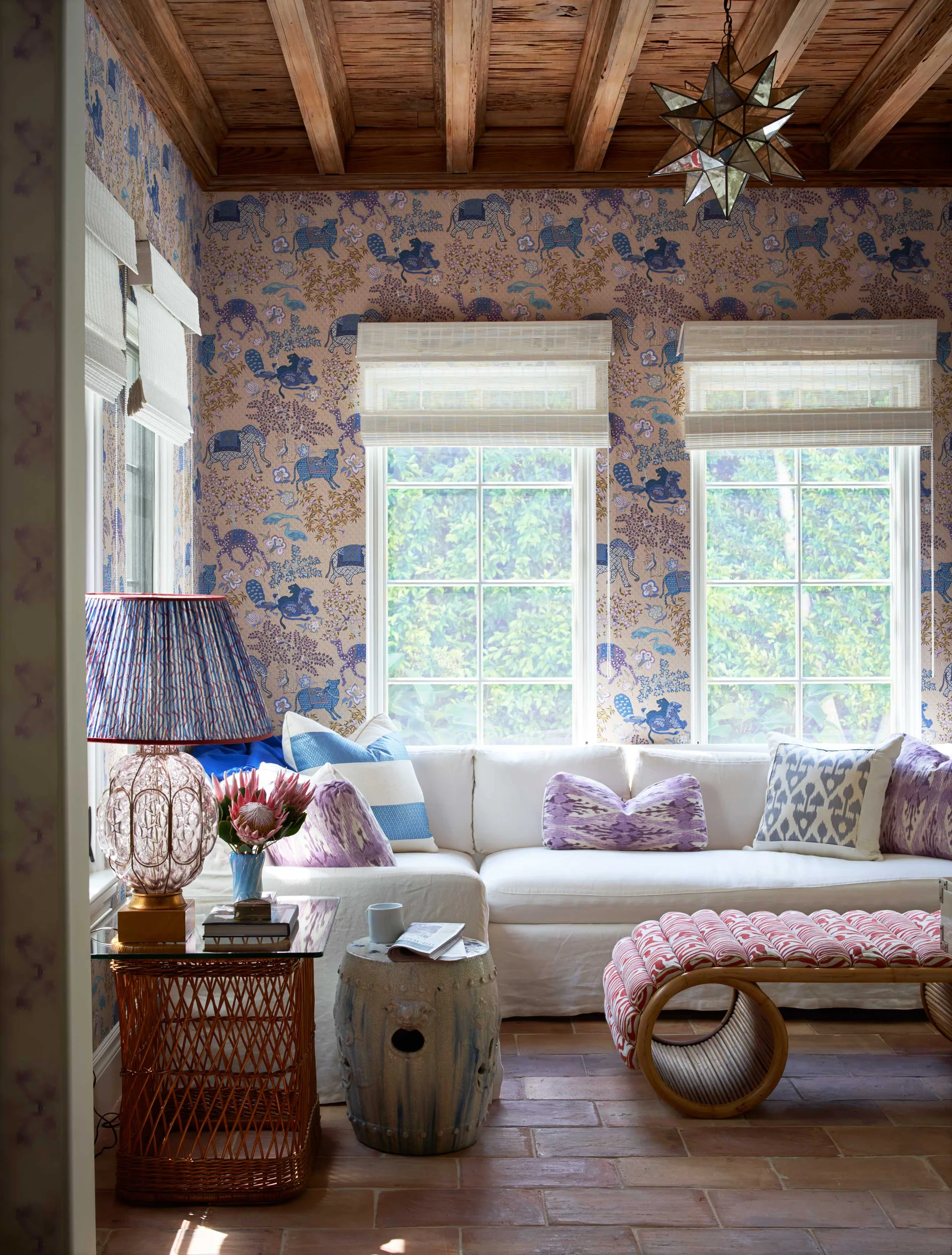 A cozy living room with animal patterned wallpaper, a white sectional sofa, three windows with roman shades, and a rustic wood plank ceiling.