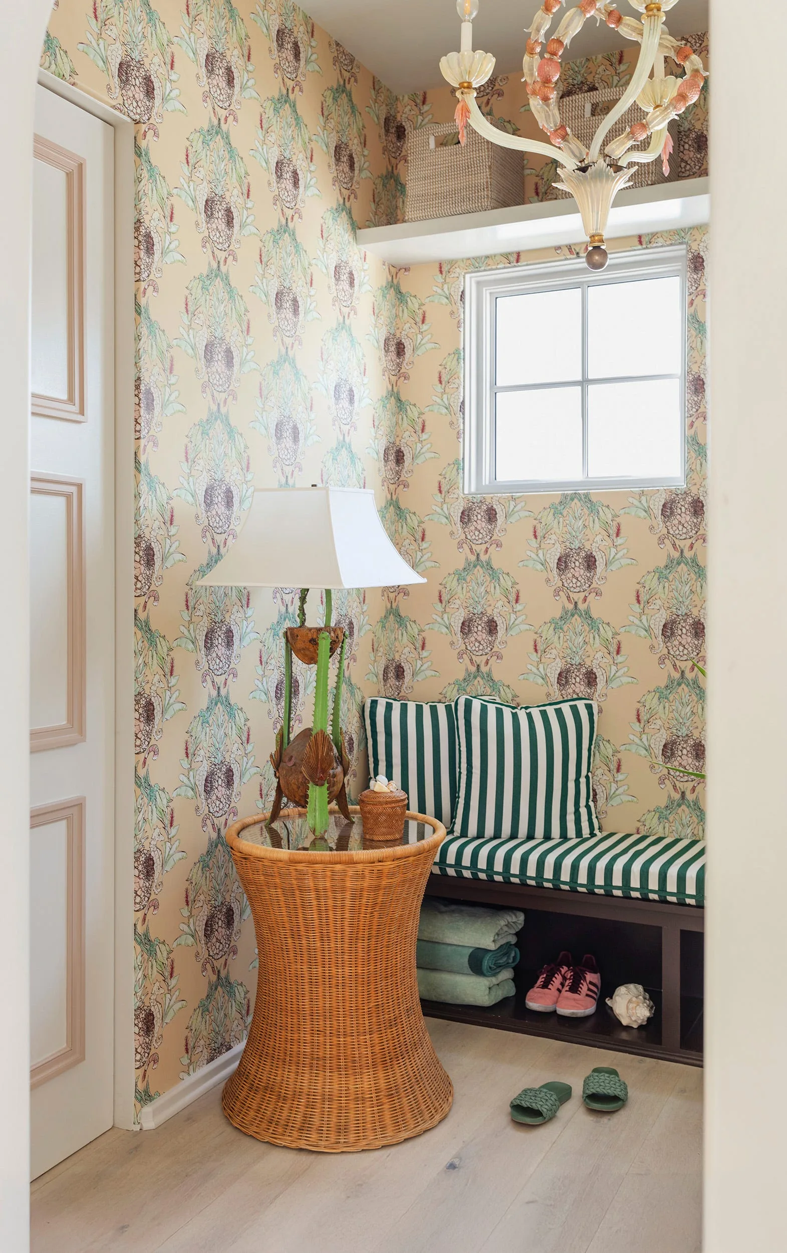 A small mudroom alcove features pineapple-patterned wallpaper, a dark wood storage bench with striped green cushions, and a wicker side table.