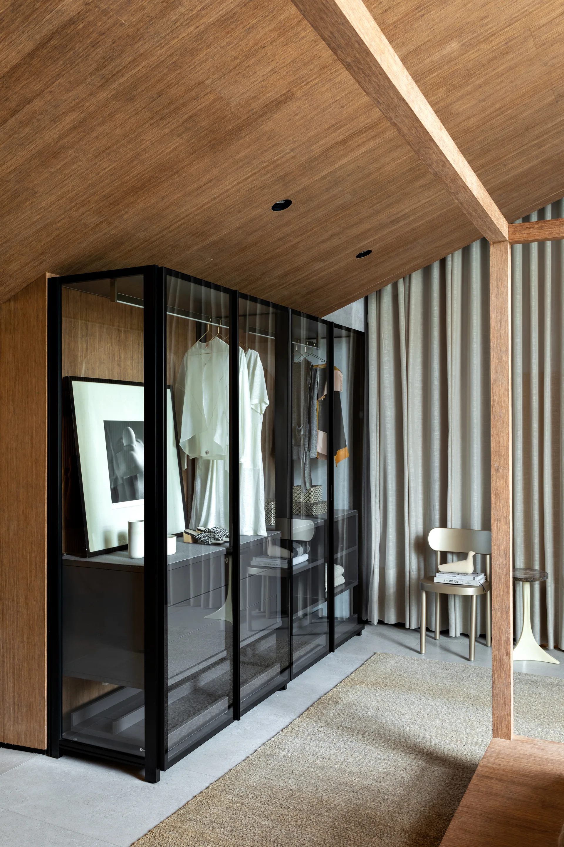 A modern dressing area features a black-framed glass wardrobe system against a sloping wood ceiling and floor-to-ceiling beige curtains.