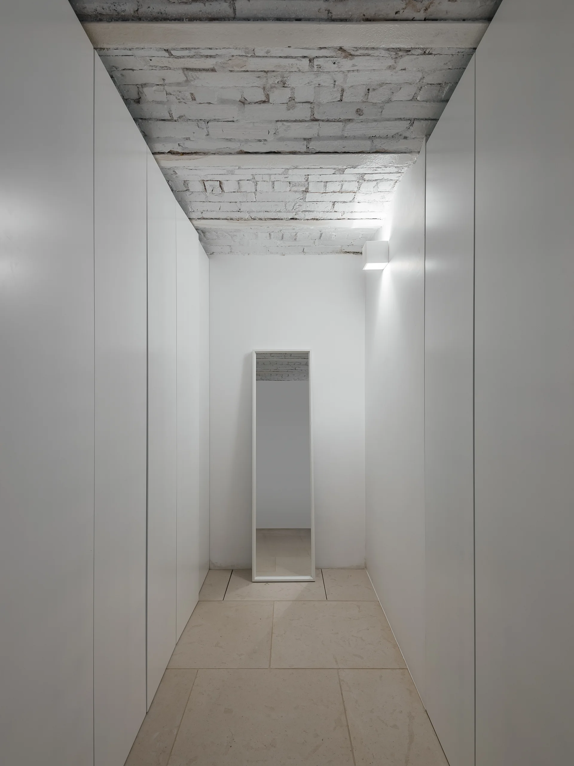 A narrow walk-in closet hallway features floor-to-ceiling white seamless wardrobes, a freestanding mirror, and an exposed white-painted brick ceiling.
