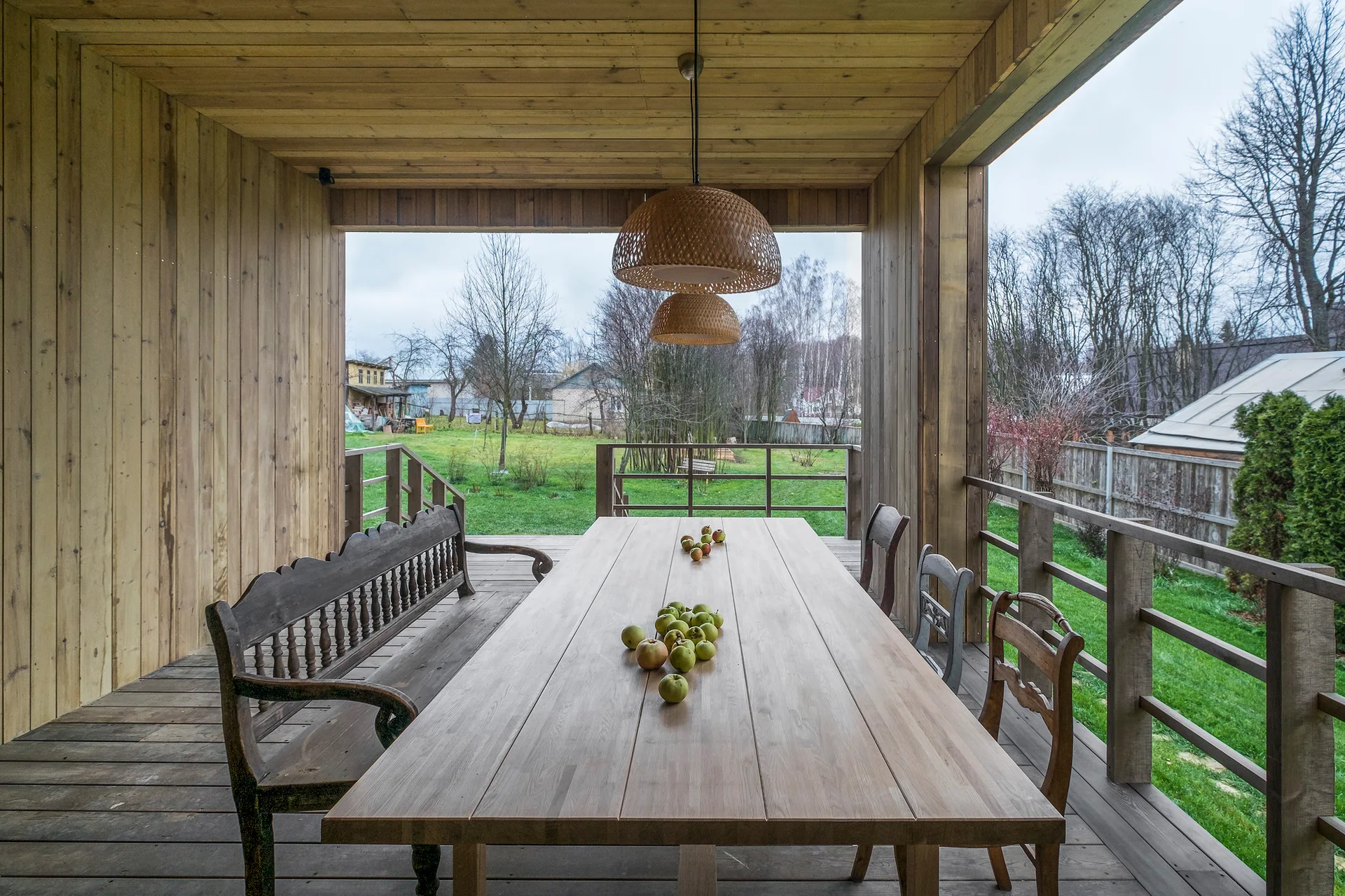 A long wooden dining table is set on a covered porch with matching wood plank walls, floor, and ceiling, featuring woven pendant lights.