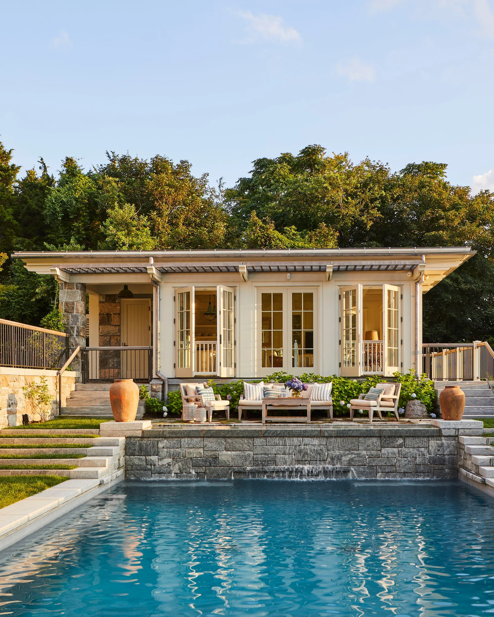 A rectangular swimming pool sits below an elevated stone patio with a cream pool house, outdoor furniture, and a built-in waterfall feature.