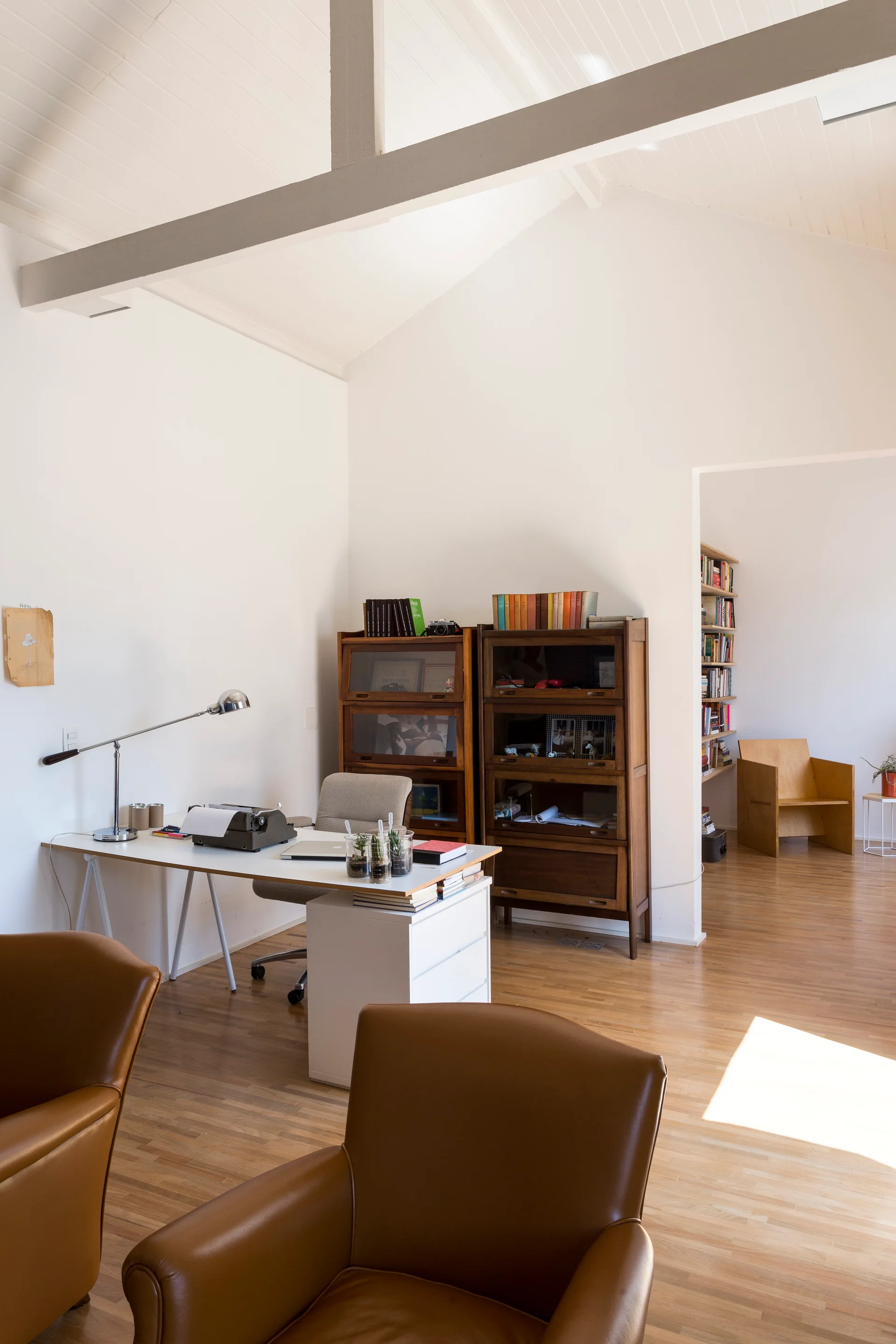 A bright home office features a white writing desk, glass-front wooden bookcases, and tan leather armchairs under a vaulted ceiling with an exposed beam.