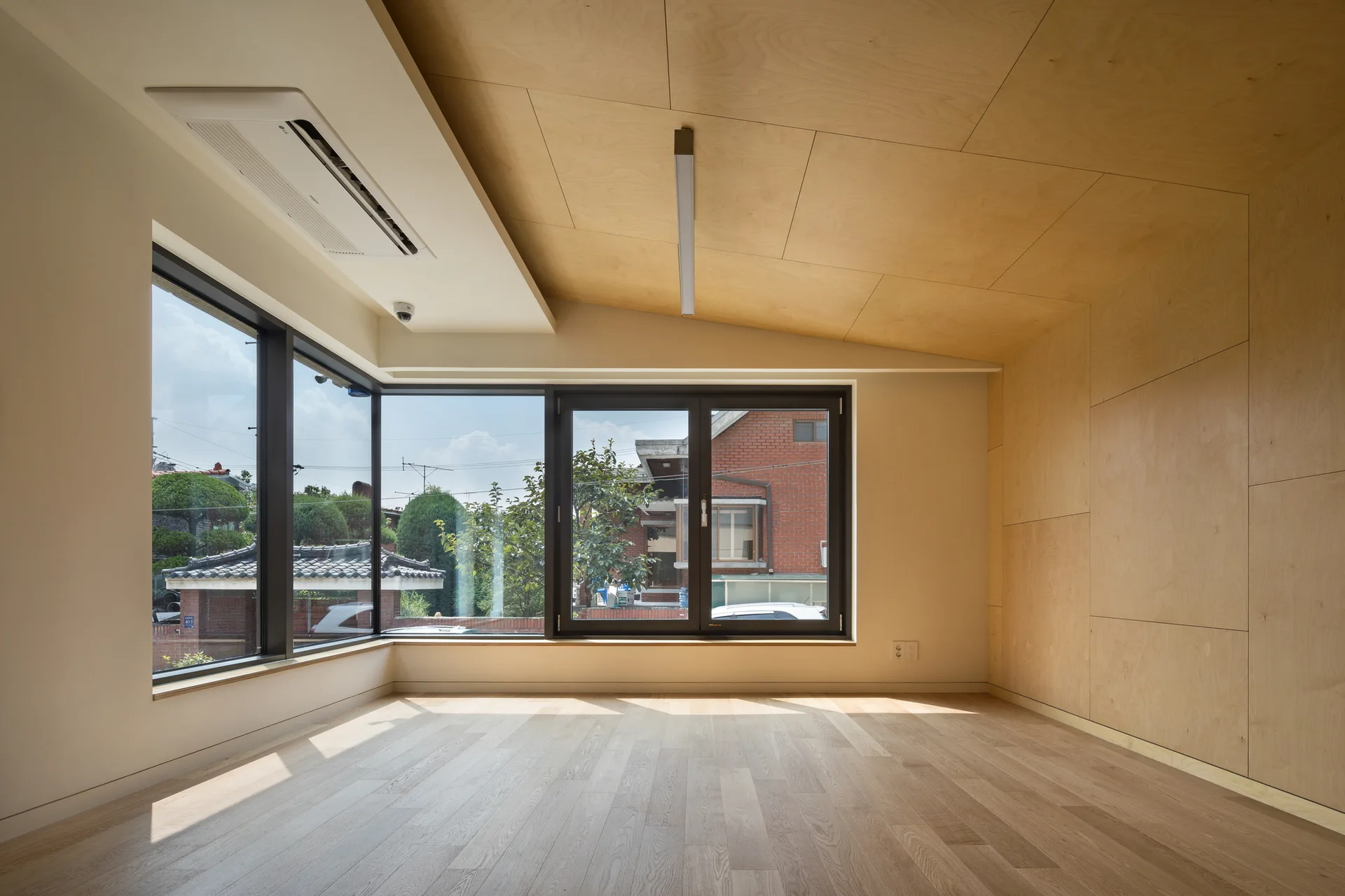 An empty minimalist room featuring light wood floors, a sloped plywood ceiling and wall, and a large black-framed corner window.