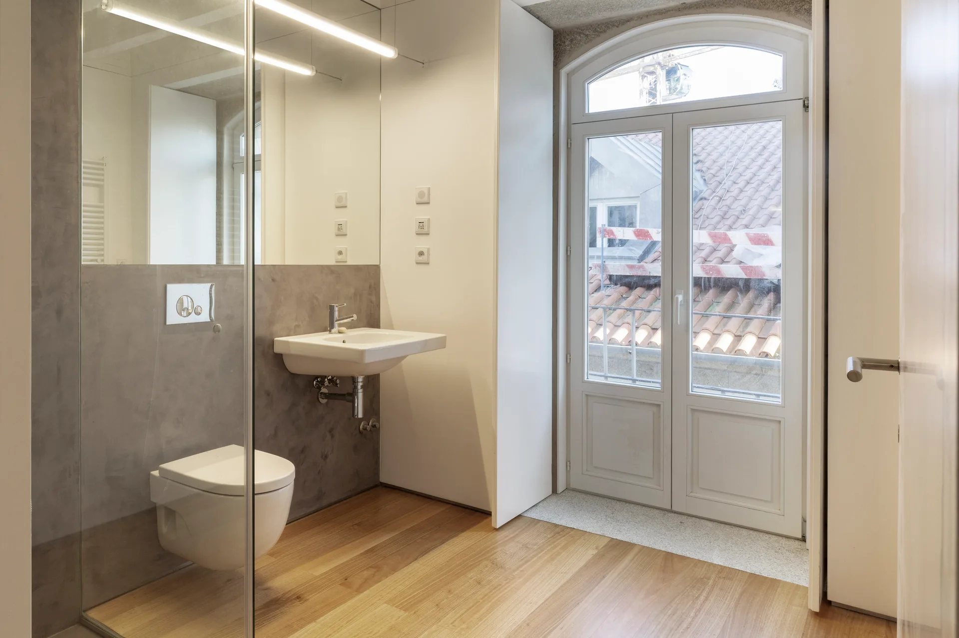 A minimalist bathroom features wood floors, grey concrete-like walls, a wall-mounted toilet and sink, and a large arched glass balcony door.