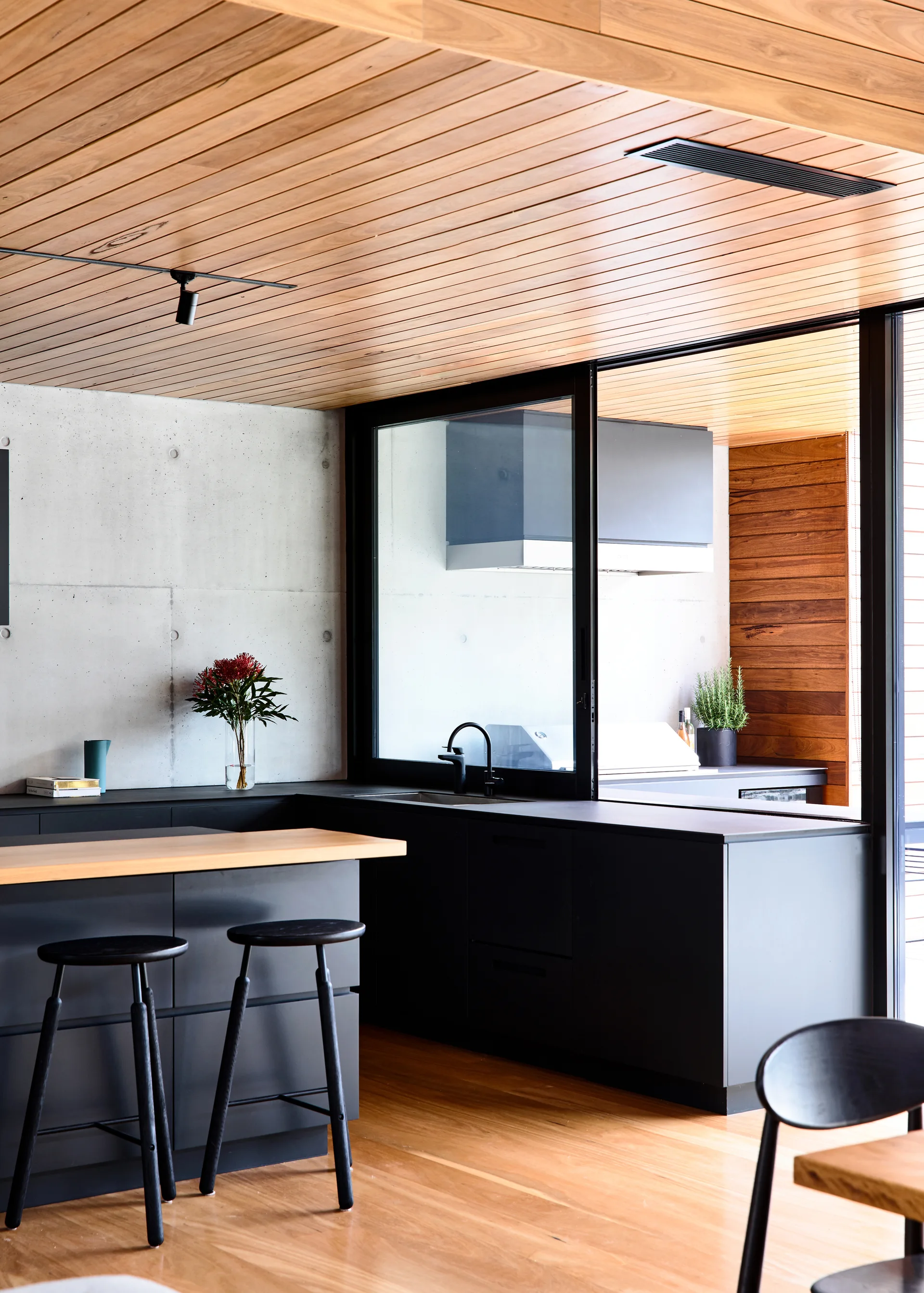 A modern kitchen features black cabinetry, a wood-topped island with black stools, a concrete wall, and a slatted timber ceiling.