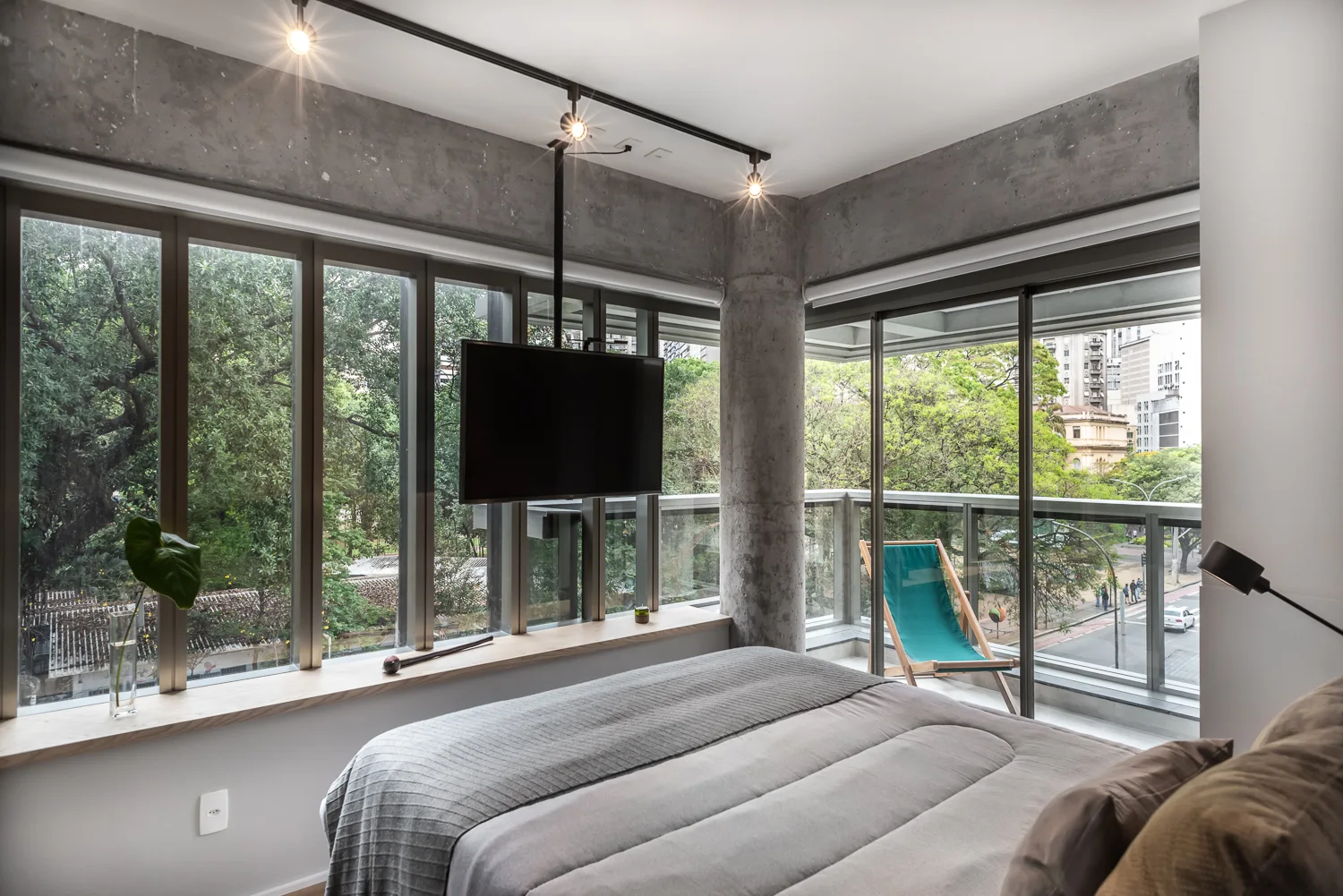 A modern bedroom features a grey bed next to expansive corner windows, a raw concrete pillar, ceiling track lighting, and a view of green trees.