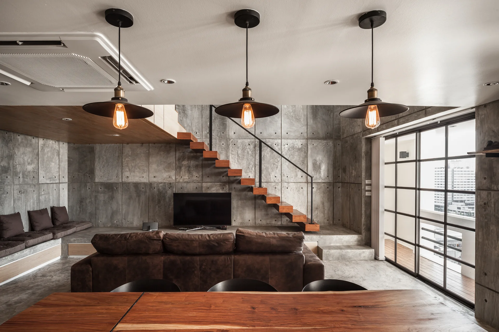 An industrial living room features a brown leather sofa, a floating wooden staircase against a concrete panel wall, and three pendant lights hanging above.