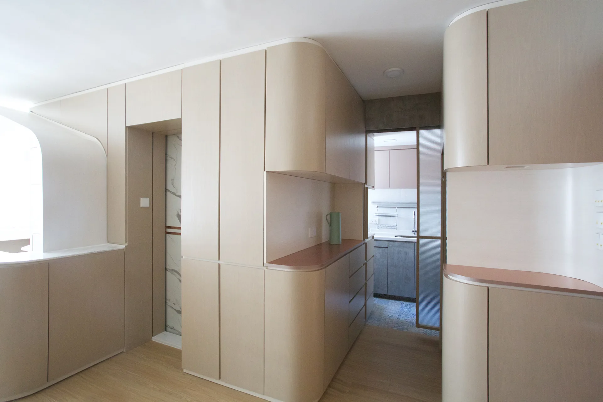 Large custom beige cabinetry with rounded corners lines a hallway leading to a kitchen with a fluted glass sliding door.
