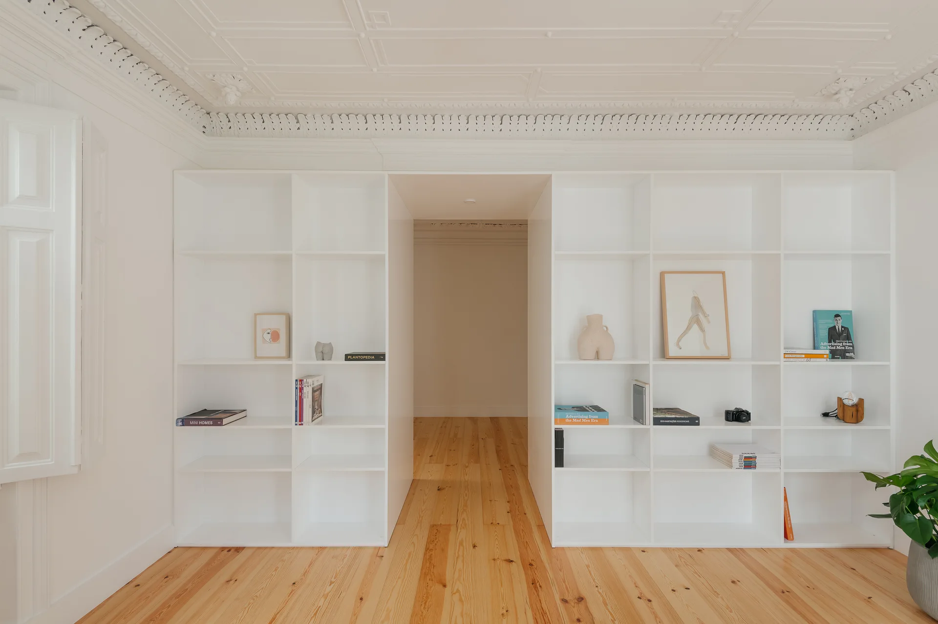 White built-in shelving units flank a central open doorway in a room with light hardwood floors and detailed ornate ceiling molding.