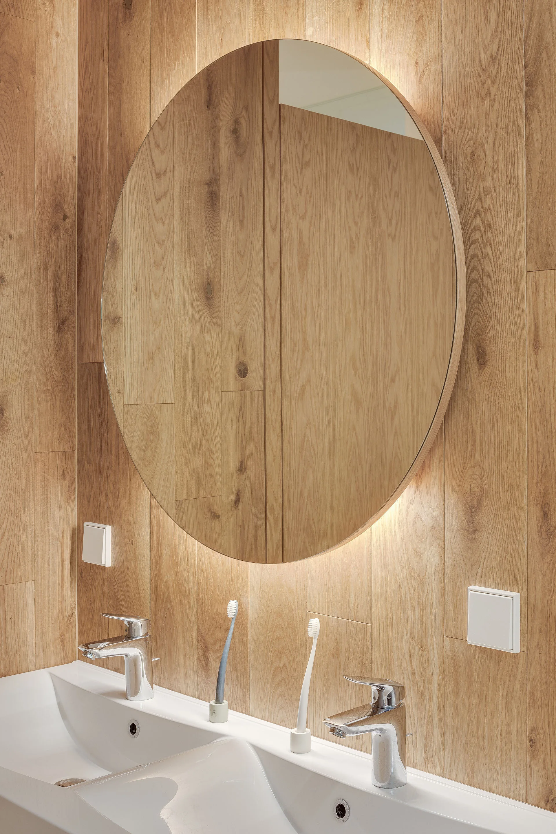 A modern bathroom vanity features a double white sink with chrome faucets and a large round backlit mirror mounted on a vertical wood paneled wall.