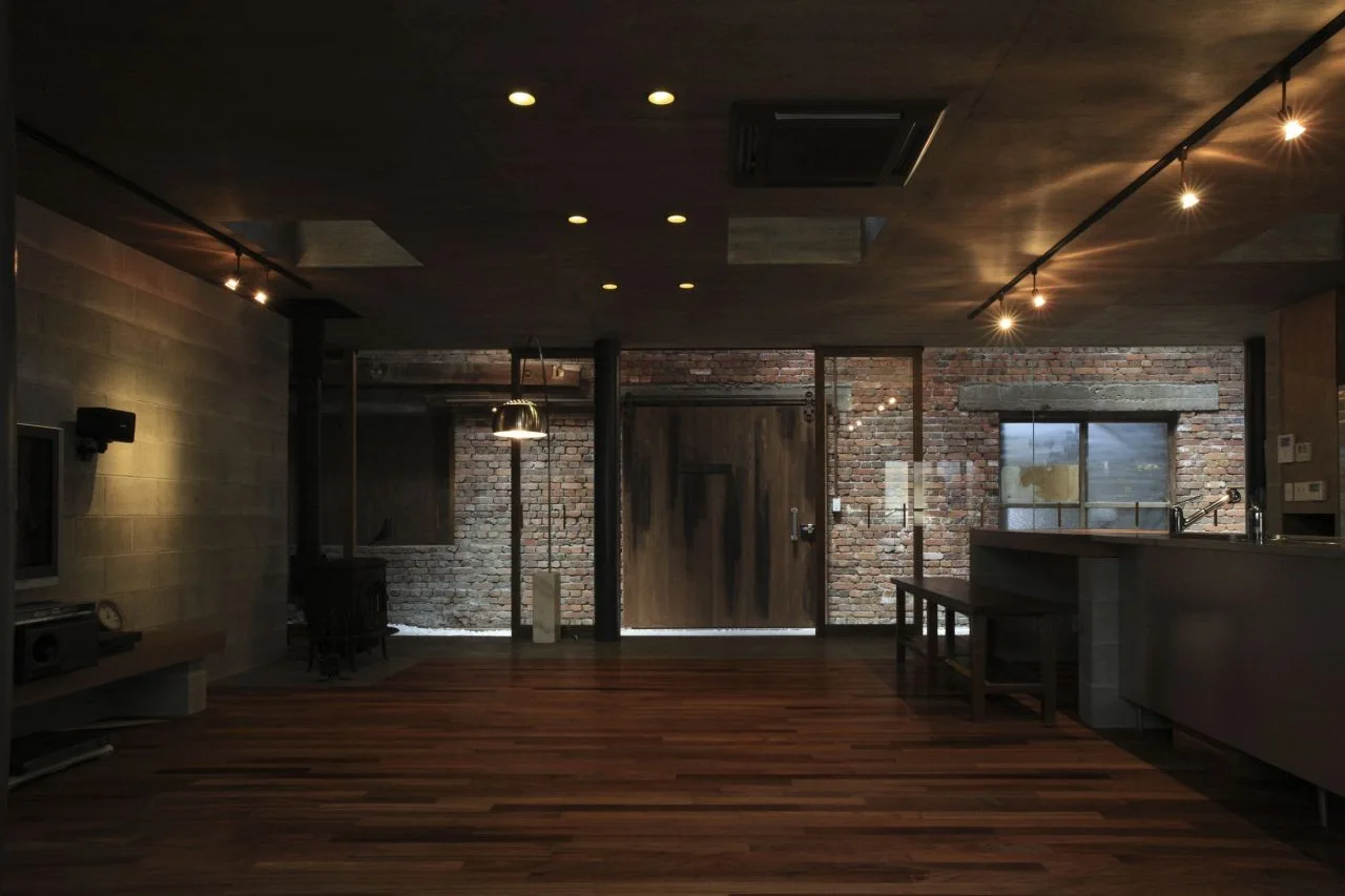 An open industrial living area features a dark ceiling with spotlights, a rustic brick wall backdrop, and dark hardwood floors.
