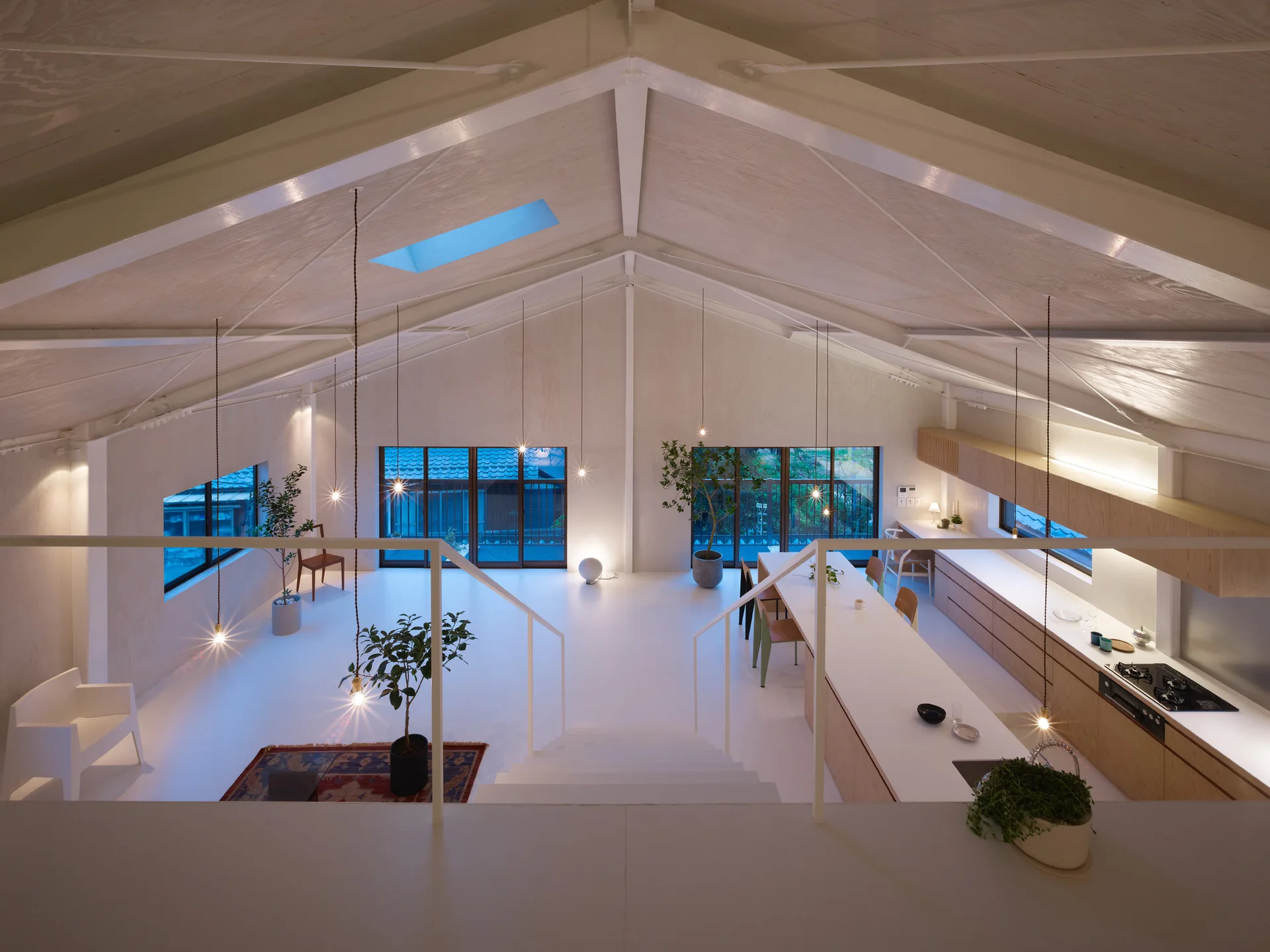 An open-plan loft features a white gabled ceiling, a long wood-toned kitchen island, and multiple bare-bulb pendant lights hanging on long cords.