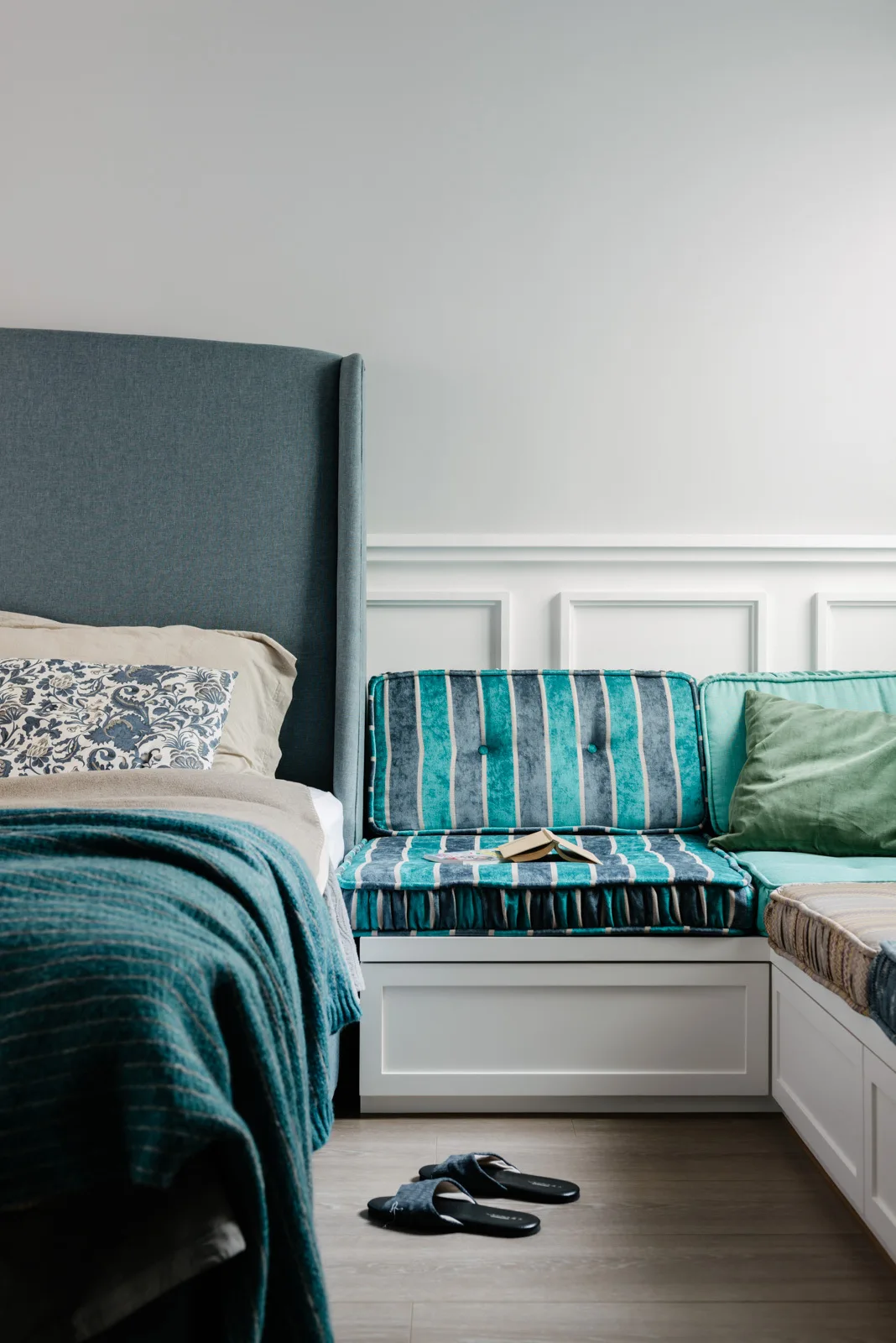 A bedroom corner features a tall blue headboard beside a white built-in bench with striped teal velvet cushions and white wainscoting on the wall.