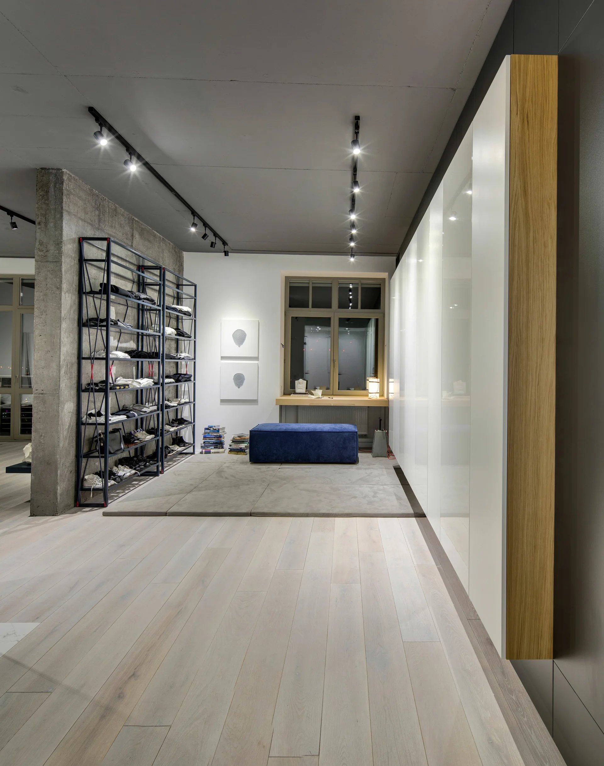 A modern dressing room features glossy white floor-to-ceiling wardrobes, a dark blue bench, an open metal shelving unit, and an exposed concrete pillar.
