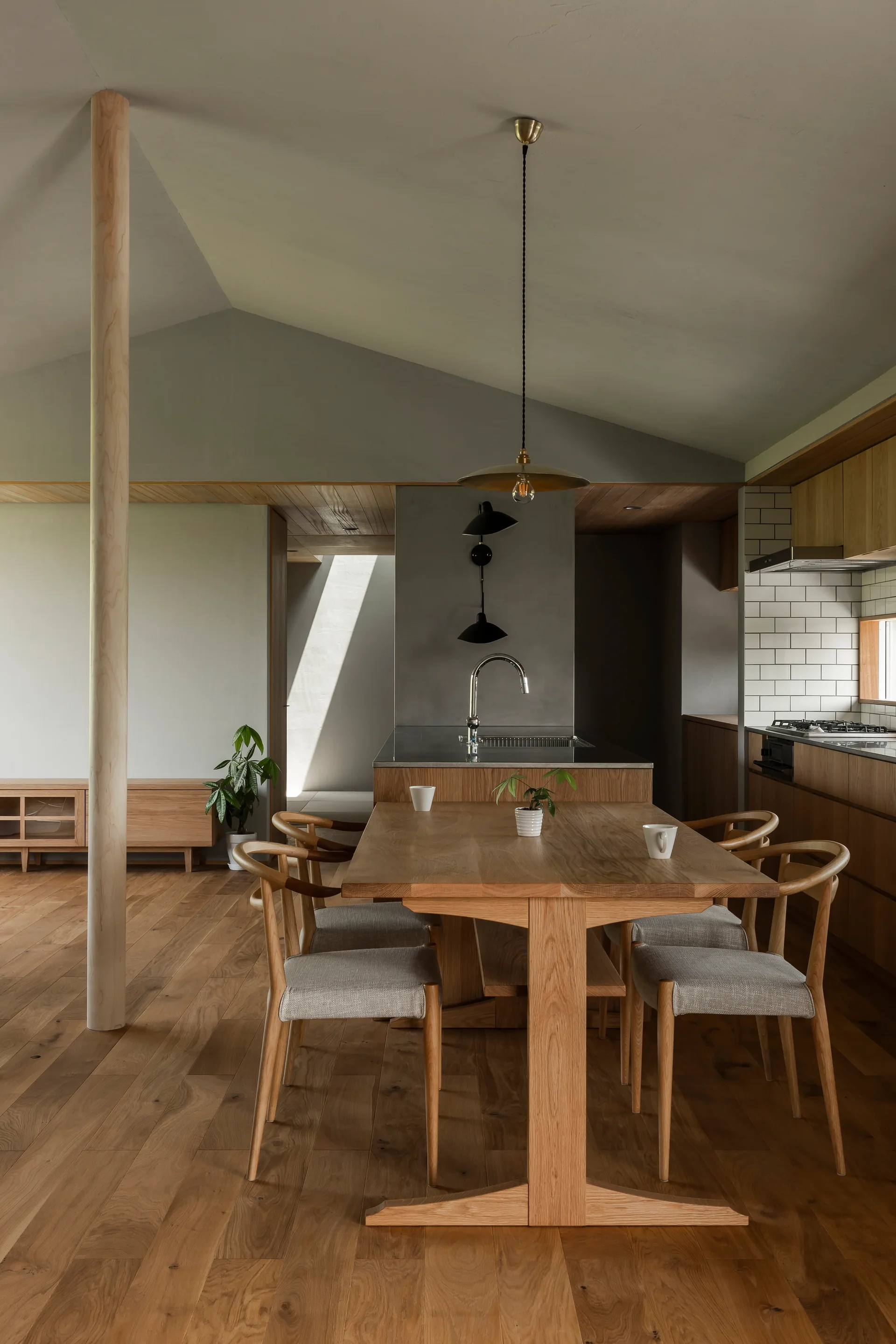 A minimalist kitchen and dining space features a light oak table, matching chairs, and an exposed round wooden column under a sloped ceiling.