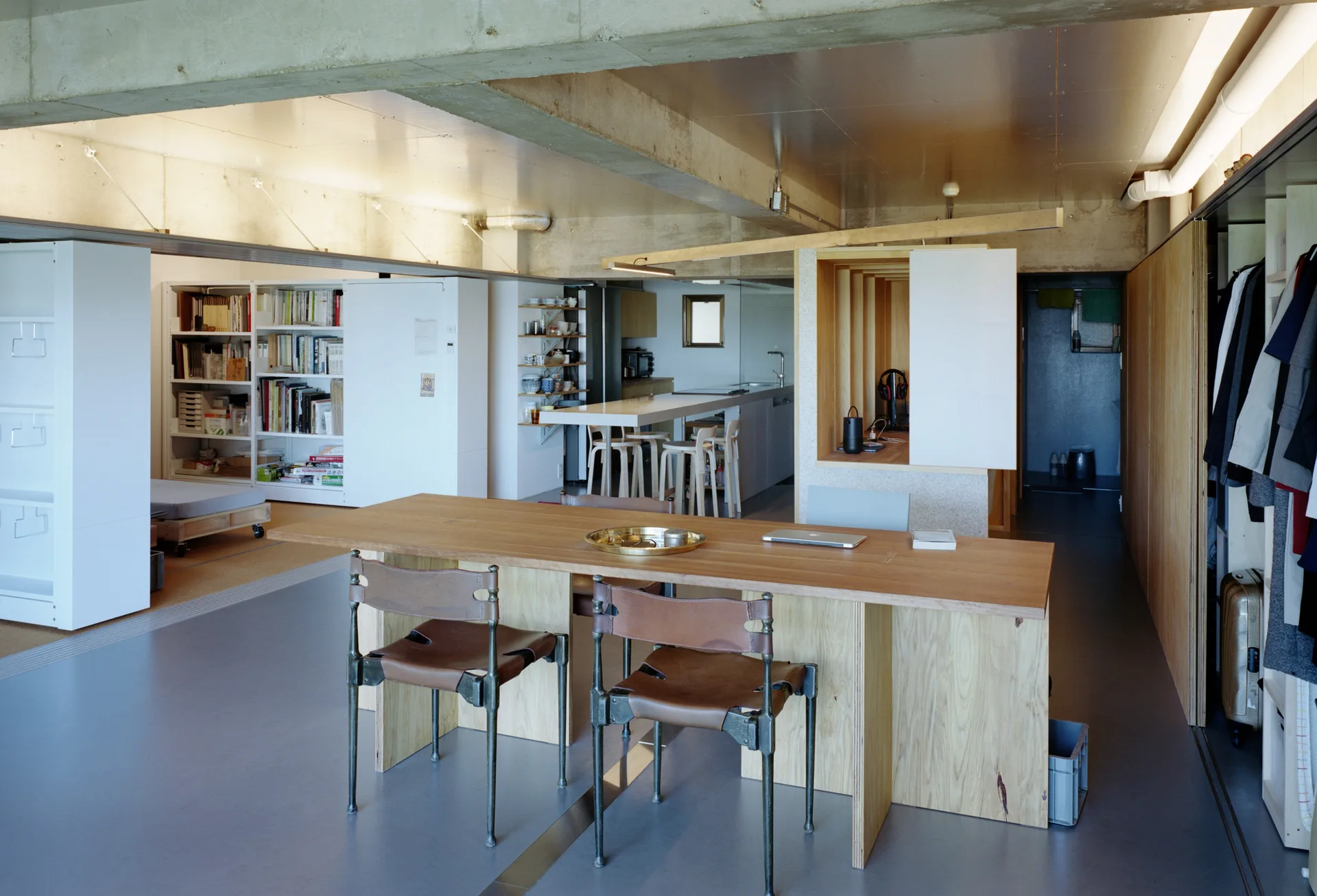 An open-plan loft space features a long plywood dining table with leather chairs under raw concrete ceiling beams.
