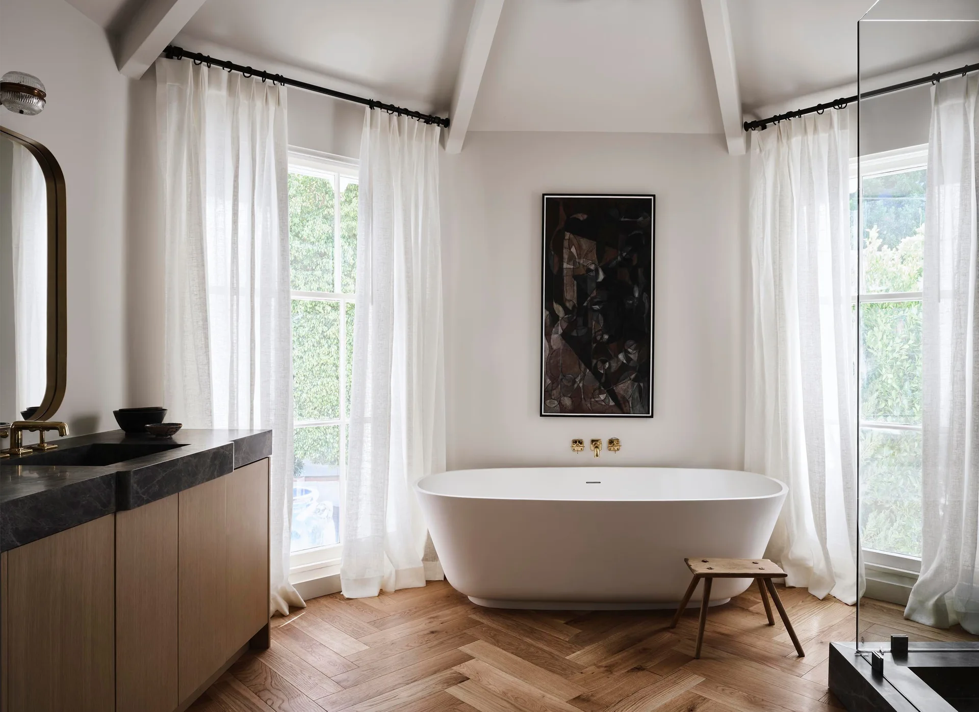 A modern bathroom features a white freestanding bathtub centered on herringbone wood floors, flanked by windows with sheer white curtains.