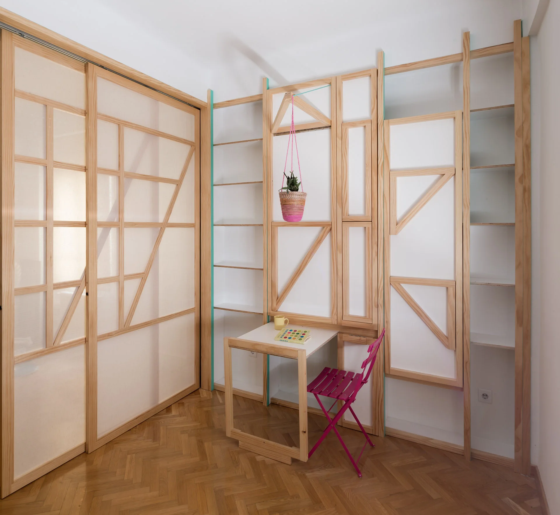 A corner home office features custom light wood built-in shelving, an integrated small desk, a bright pink folding chair, and herringbone flooring.