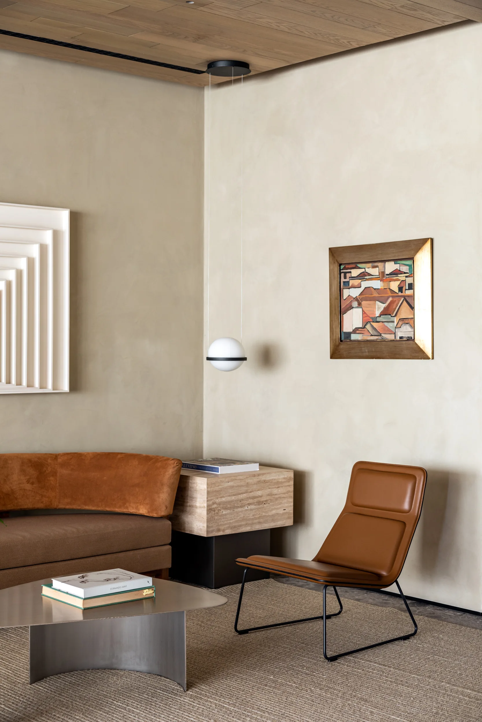 A contemporary living room featuring a curved brown sofa, a leather accent chair, a travertine side table, and smooth beige plaster walls under a wood ceiling.