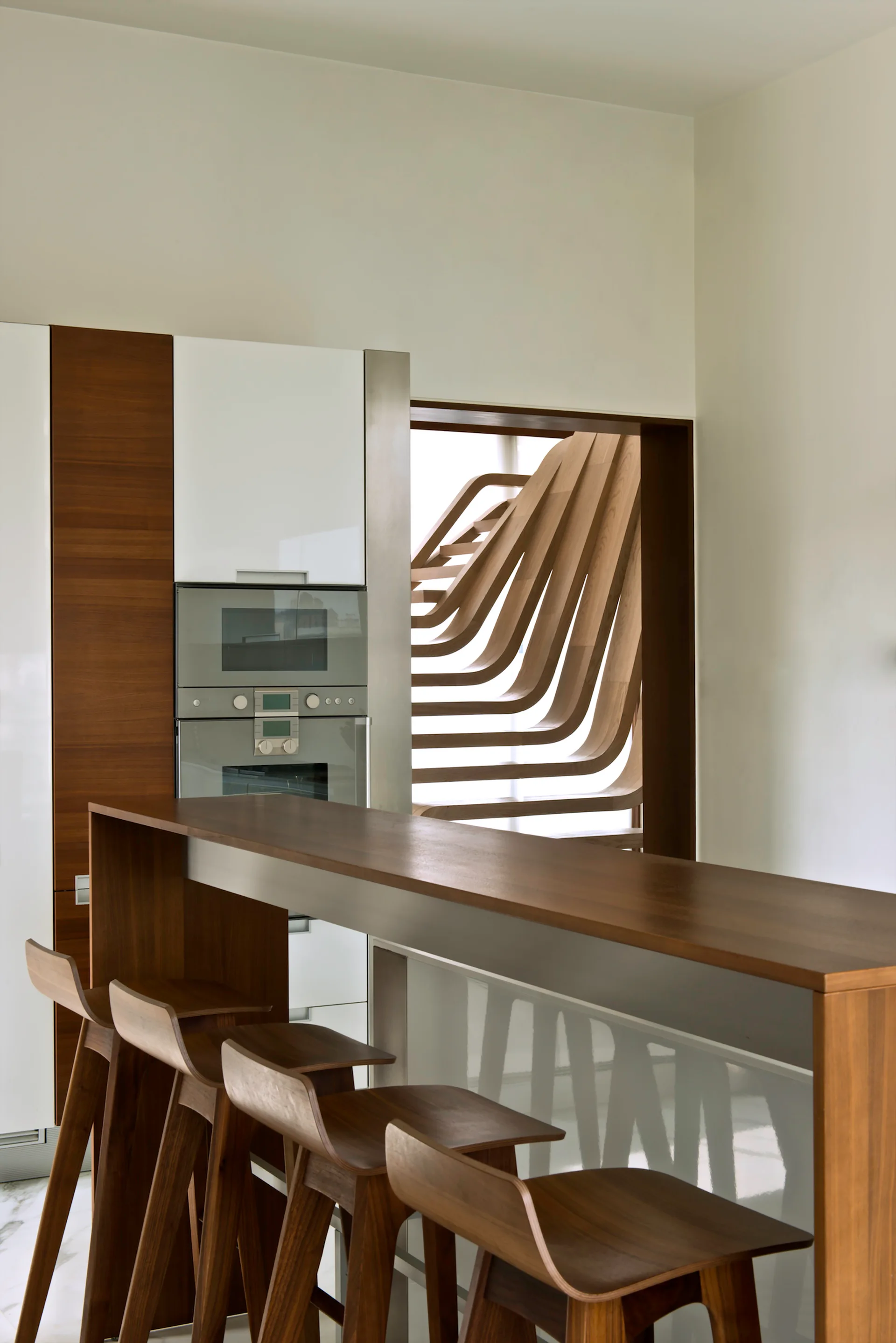 A modern kitchen with a wood-topped breakfast bar and a distinctive curved wooden staircase visible through a wall opening.