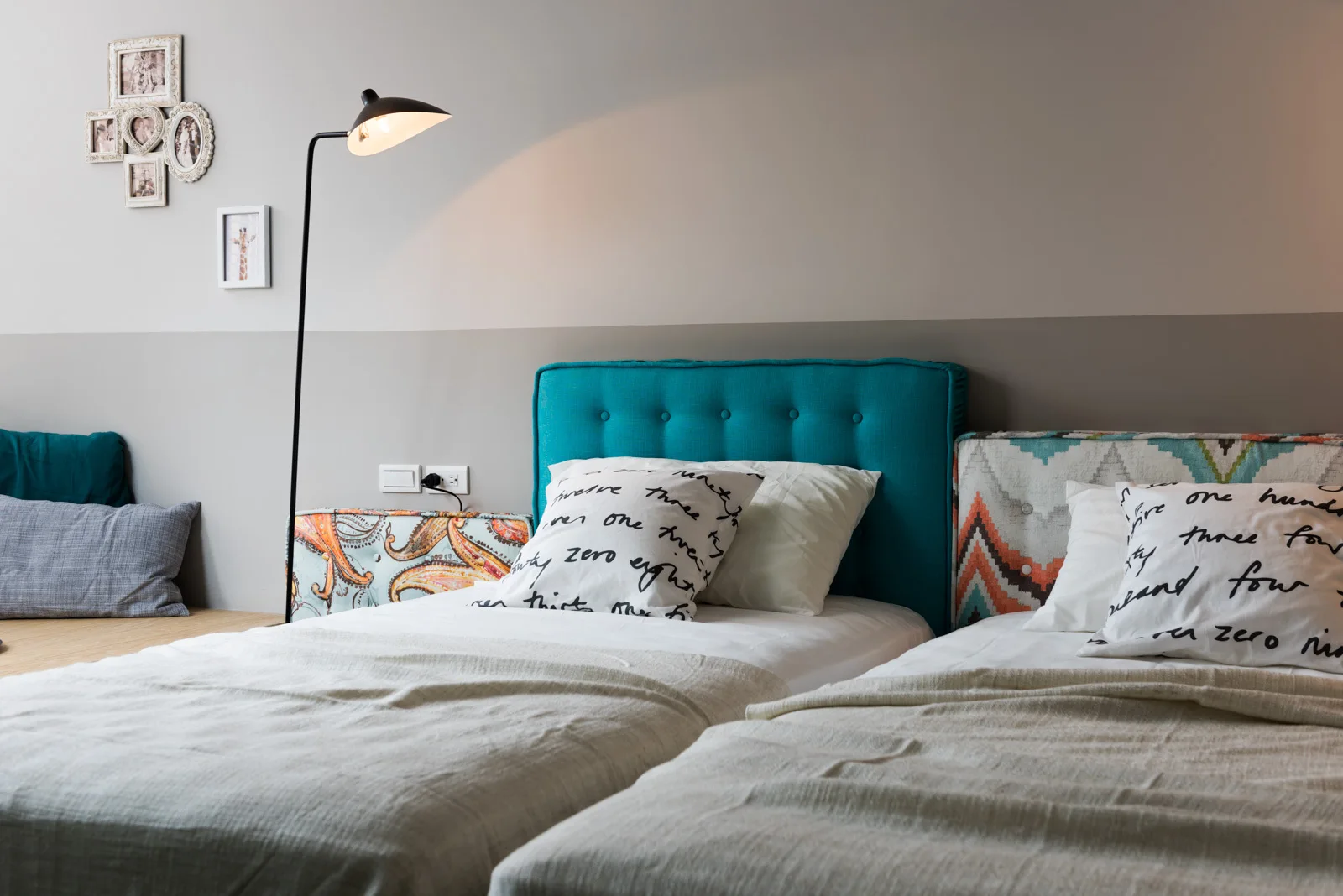 Two twin beds with different headboards stand against a two-tone grey wall in a casual bedroom next to a black floor lamp.