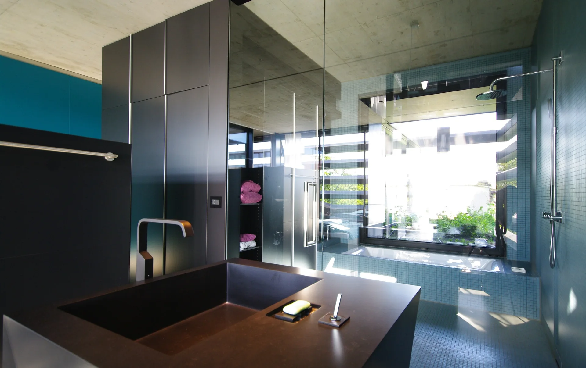 A modern bathroom features a dark vanity in the foreground, light blue mosaic tiles on the walls and floor, a built-in tub by a window, and a concrete ceiling.