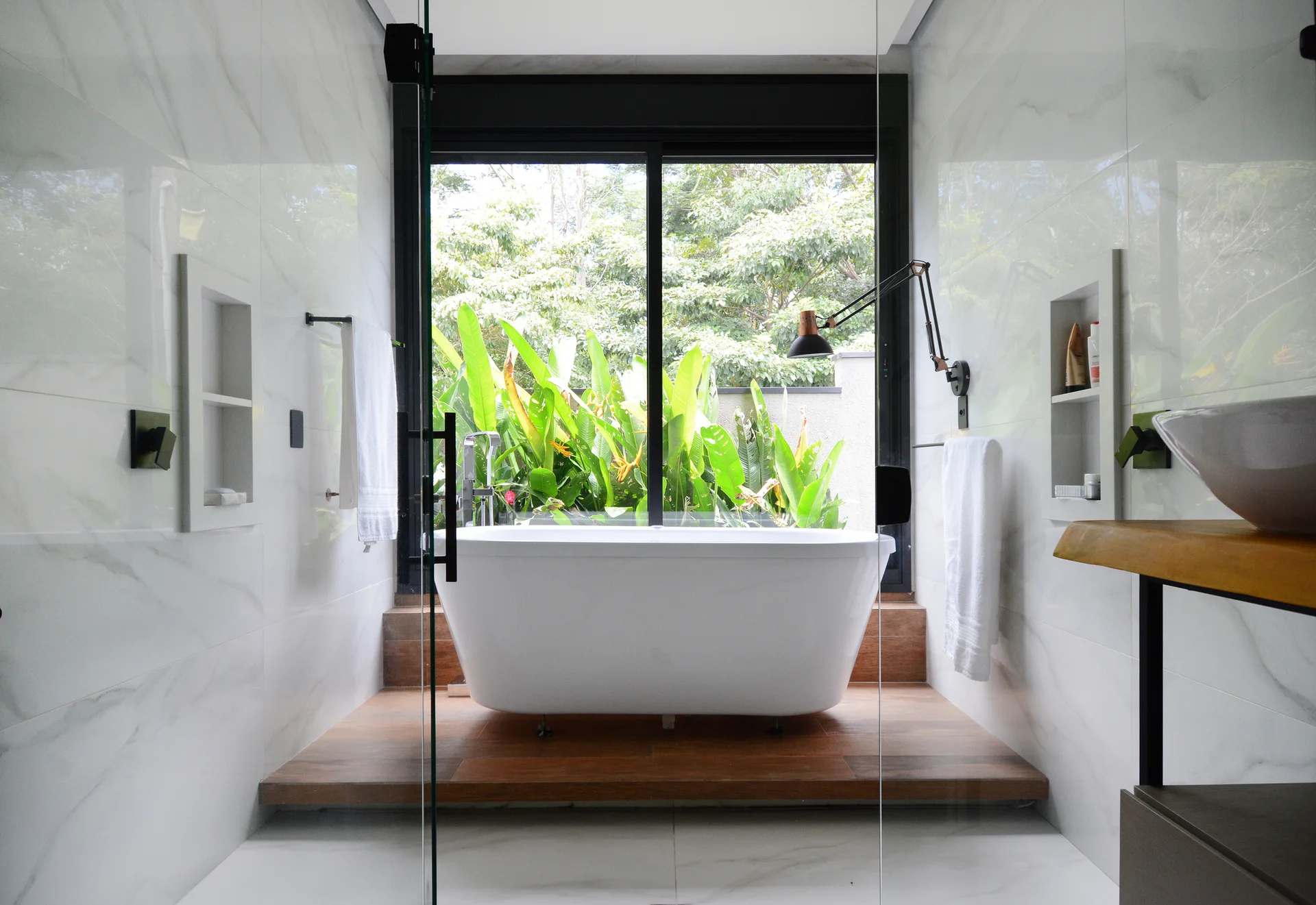 A white freestanding bathtub sits on a raised wood platform in front of a large window overlooking green foliage, surrounded by white marble-like tile walls.