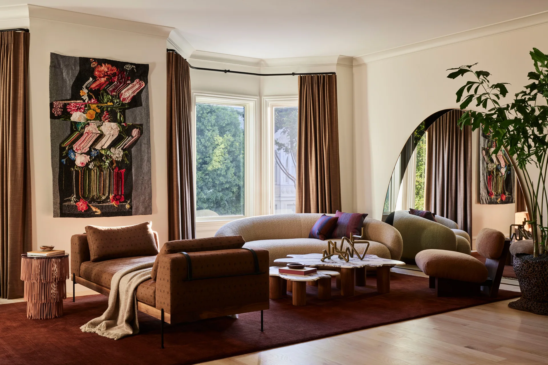 A contemporary living room features a curved cream sofa, a brown daybed, and a large arched mirror reflecting the window drapery.