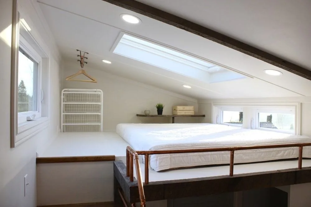 A compact loft bedroom featuring a platform bed with white bedding, a copper pipe railing, and a sloped ceiling with a skylight.