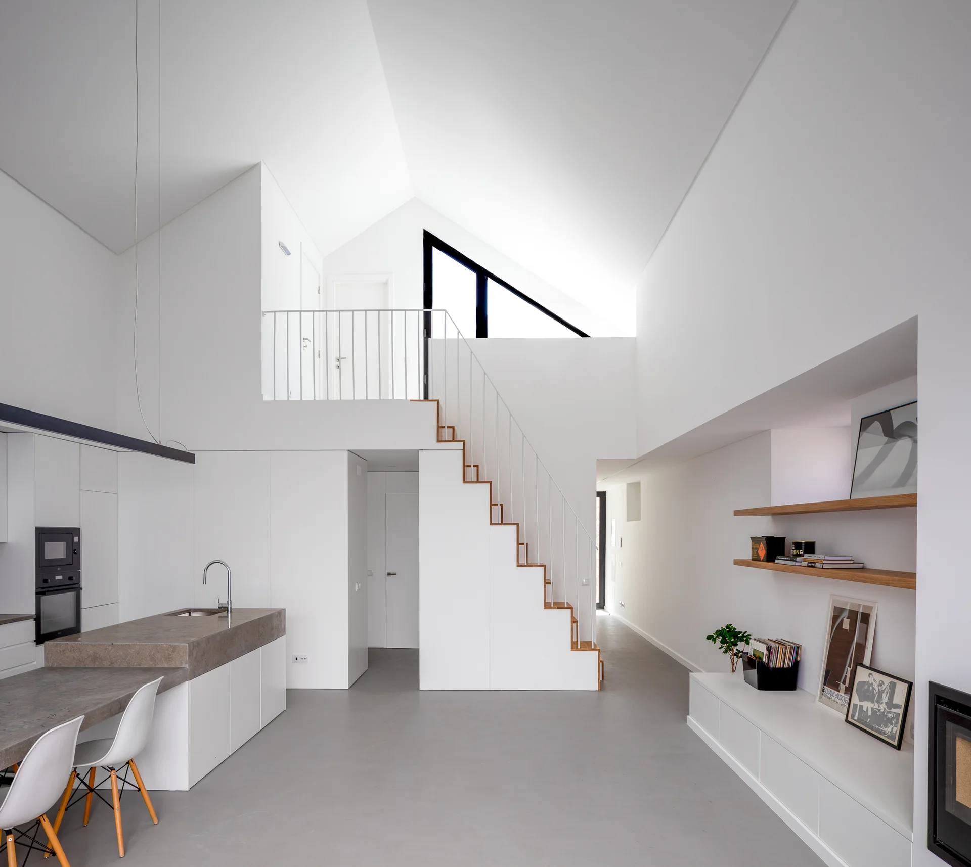 A modern minimalist interior featuring a white staircase with wood treads, a grey kitchen island, and a mezzanine under a vaulted ceiling.