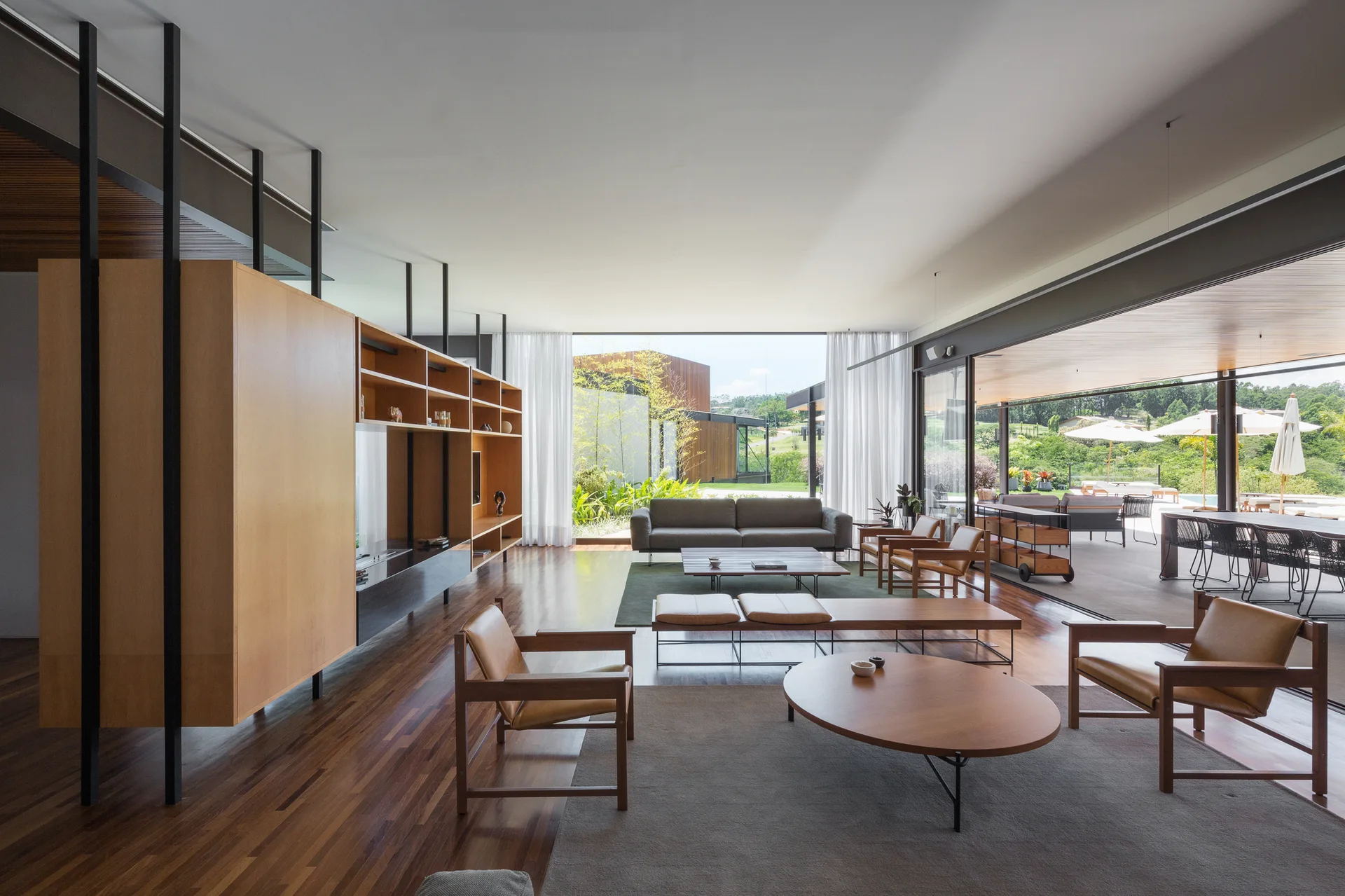 A modern open-plan living room features wood lounge chairs, a large built-in wooden shelving unit, and sliding glass doors opening to a patio.