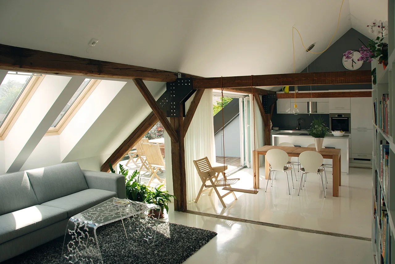 A modern attic living space features exposed wooden beams, a grey sofa, skylights, a wooden dining set, and a glossy white floor.