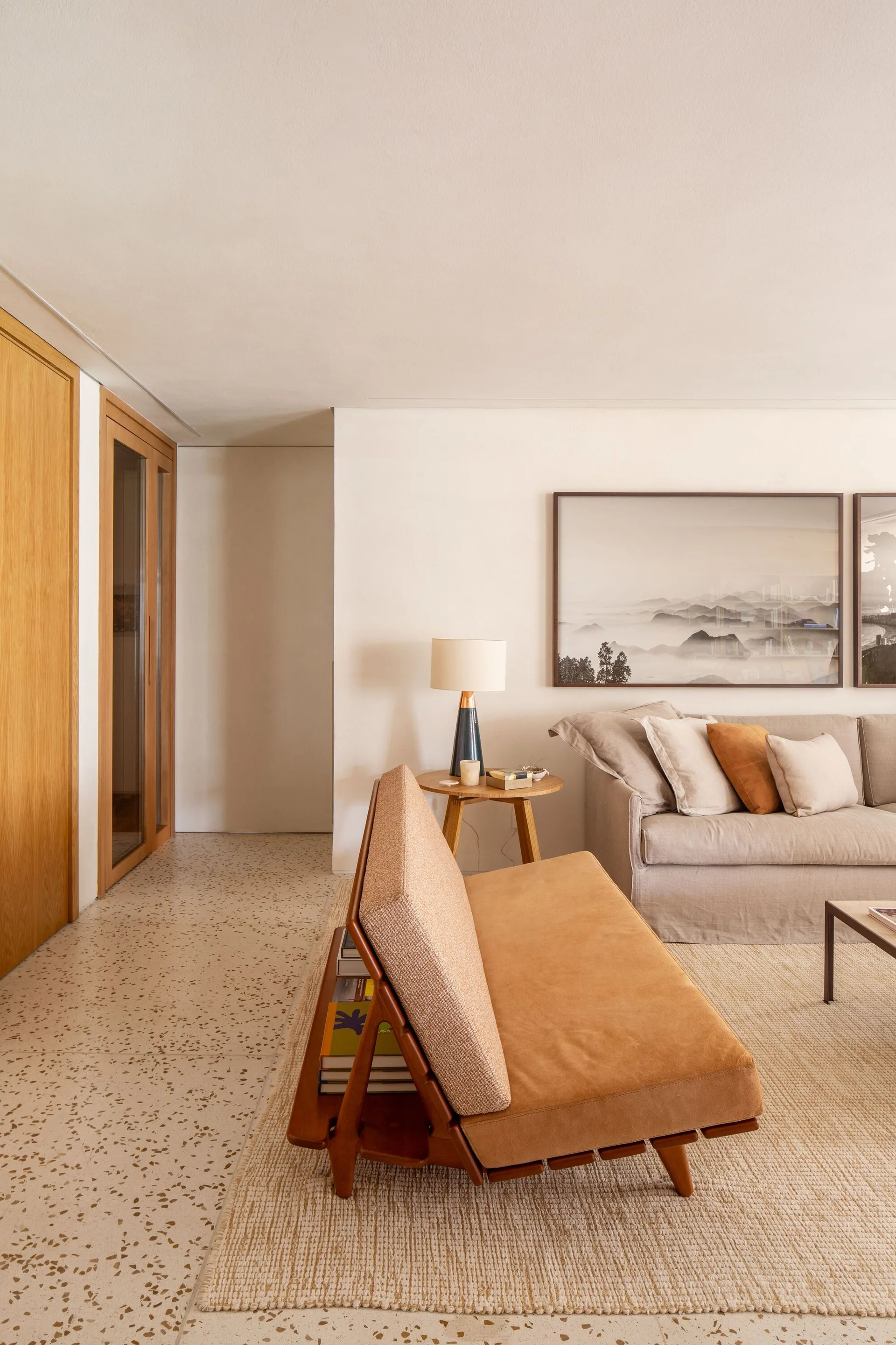 A living room features a tan wood-frame sofa, a beige linen couch, terrazzo flooring, and a large framed landscape art piece on an off-white wall.