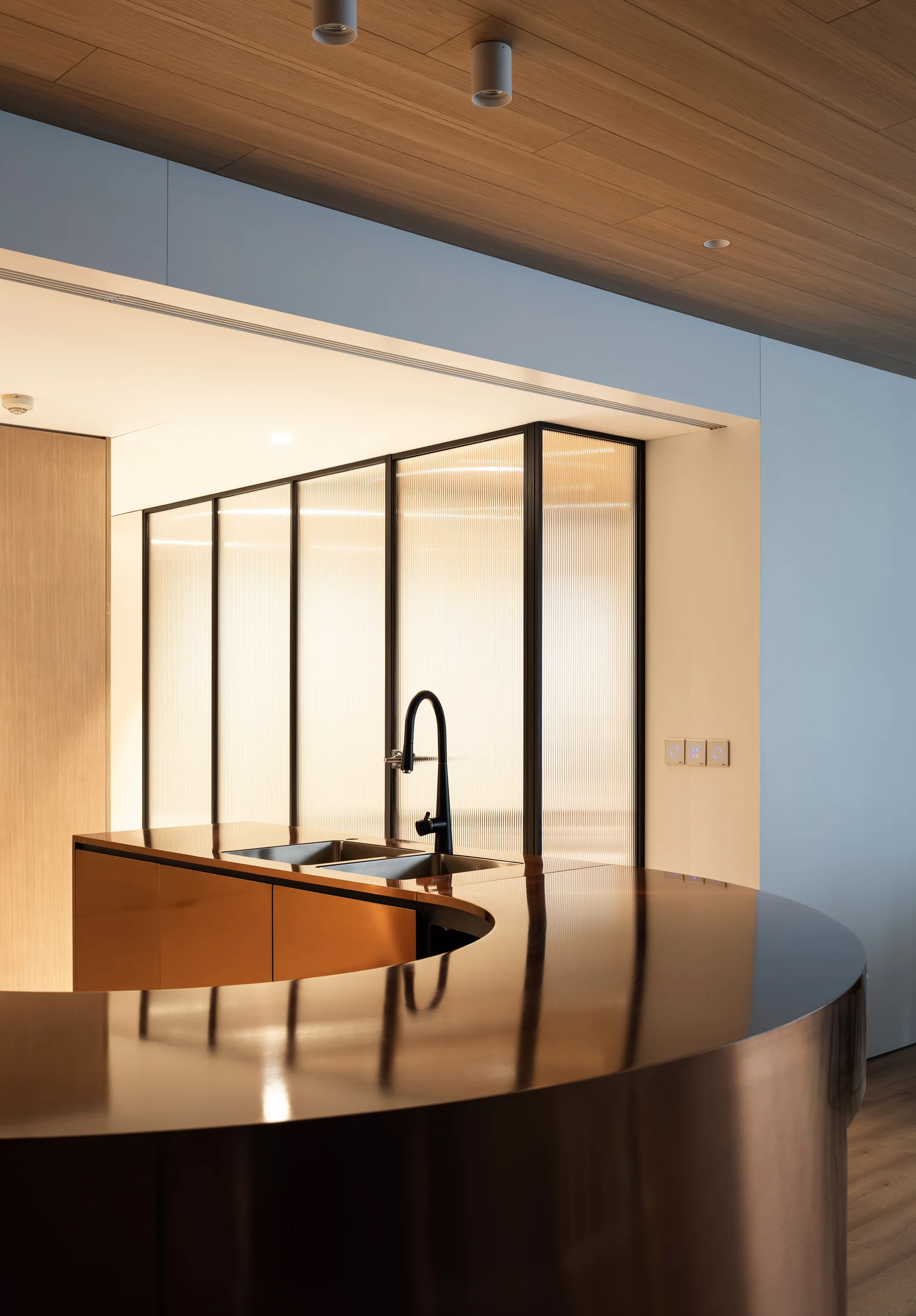 A modern kitchen featuring a curved bronze-finished island with a black faucet in front of a backlit reeded glass partition.