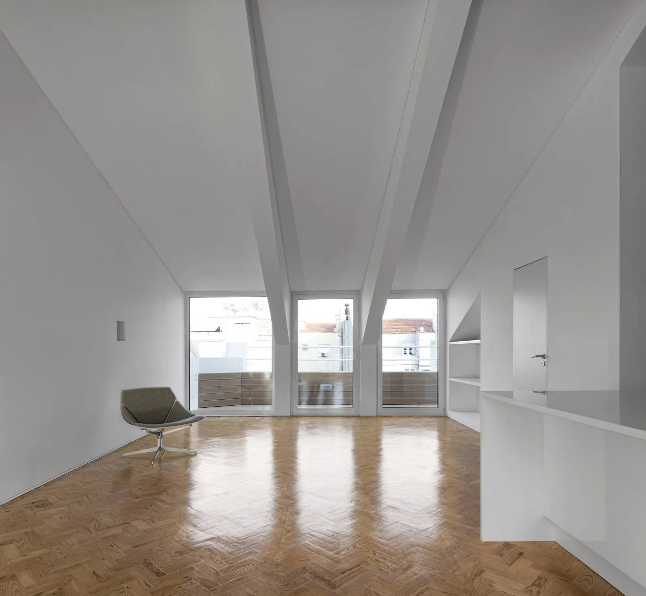 A minimalist room featuring white sloped ceilings with structural beams, a grey armchair, three tall windows, and glossy herringbone wood flooring.