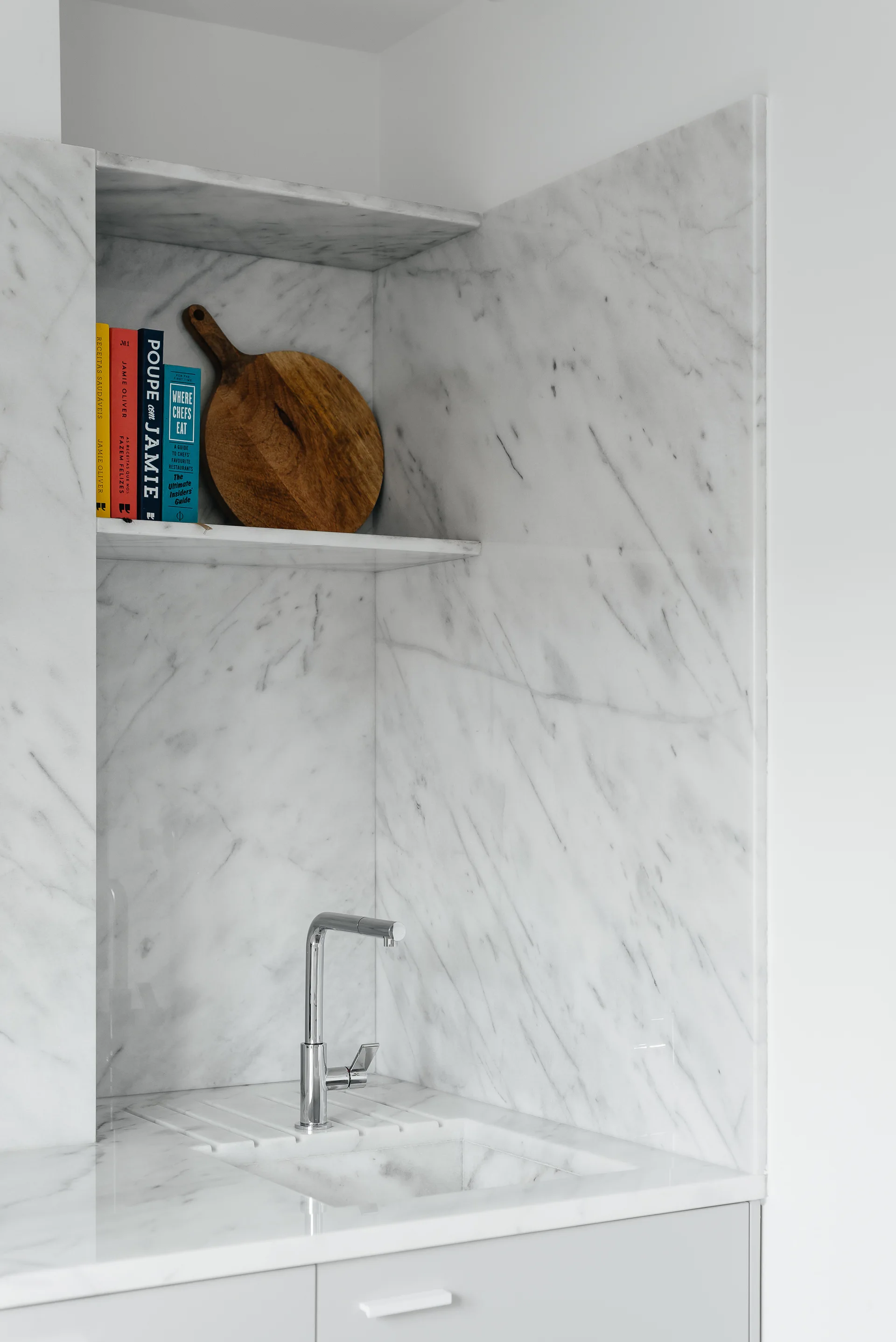 A modern kitchen featuring an integrated marble sink, a chrome faucet, and a recessed marble wall niche with decorative books and a wooden cutting board.