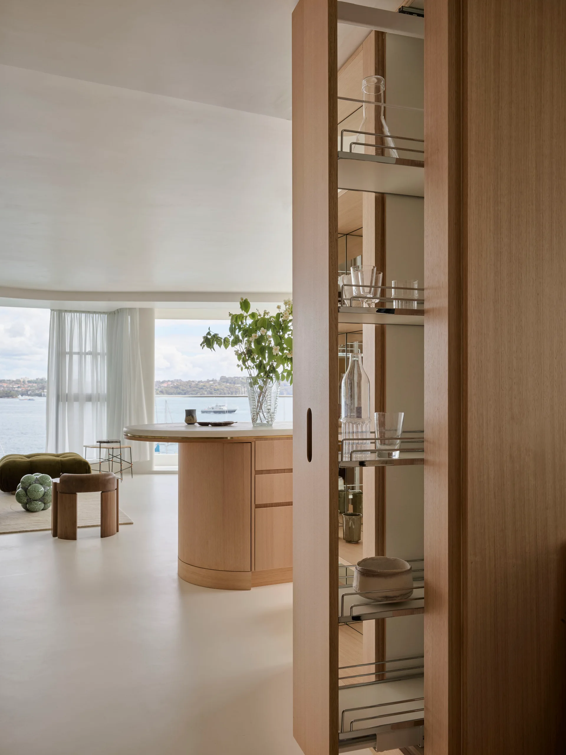 A modern kitchen features a tall wooden pull-out pantry with wire shelves next to a curved wood island overlooking a scenic water view.