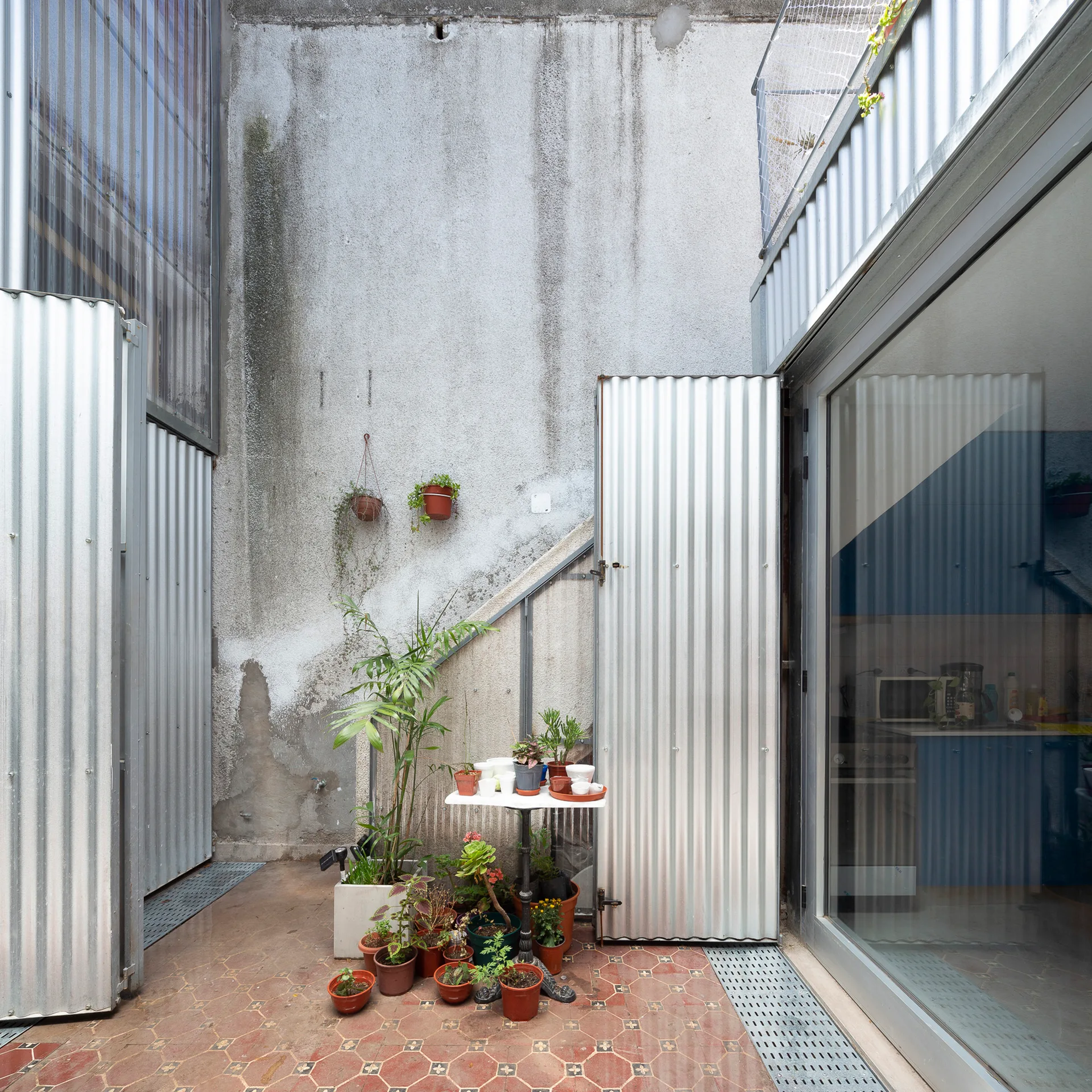 An outdoor courtyard features corrugated metal panels, a weathered concrete wall, and potted plants on patterned terracotta tiles.