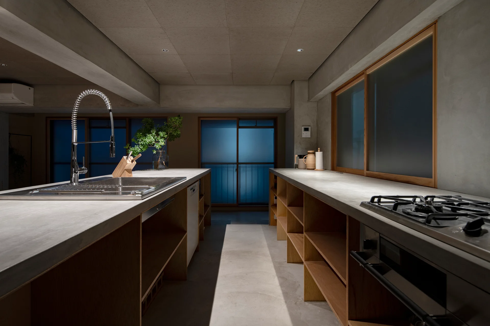 A minimalist kitchen features a concrete island with a tall chrome faucet and parallel open wood shelving under a concrete countertop.