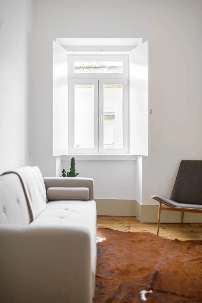 A minimalist living room with a cream sofa, dark grey lounge chair, brown animal hide rug, and a tall window with white interior shutters.