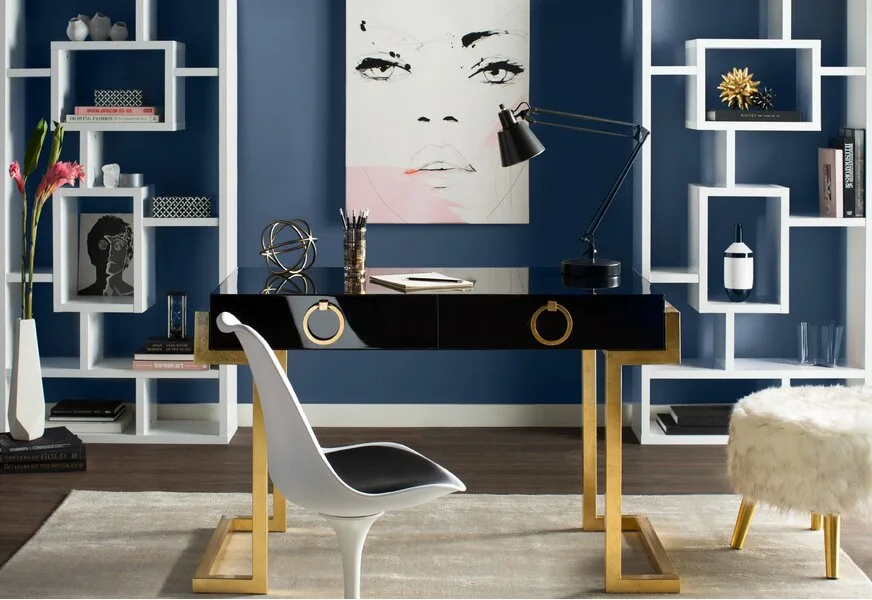 A modern home office features a glossy black desk with gold legs set against a navy blue wall between two white geometric bookcases.