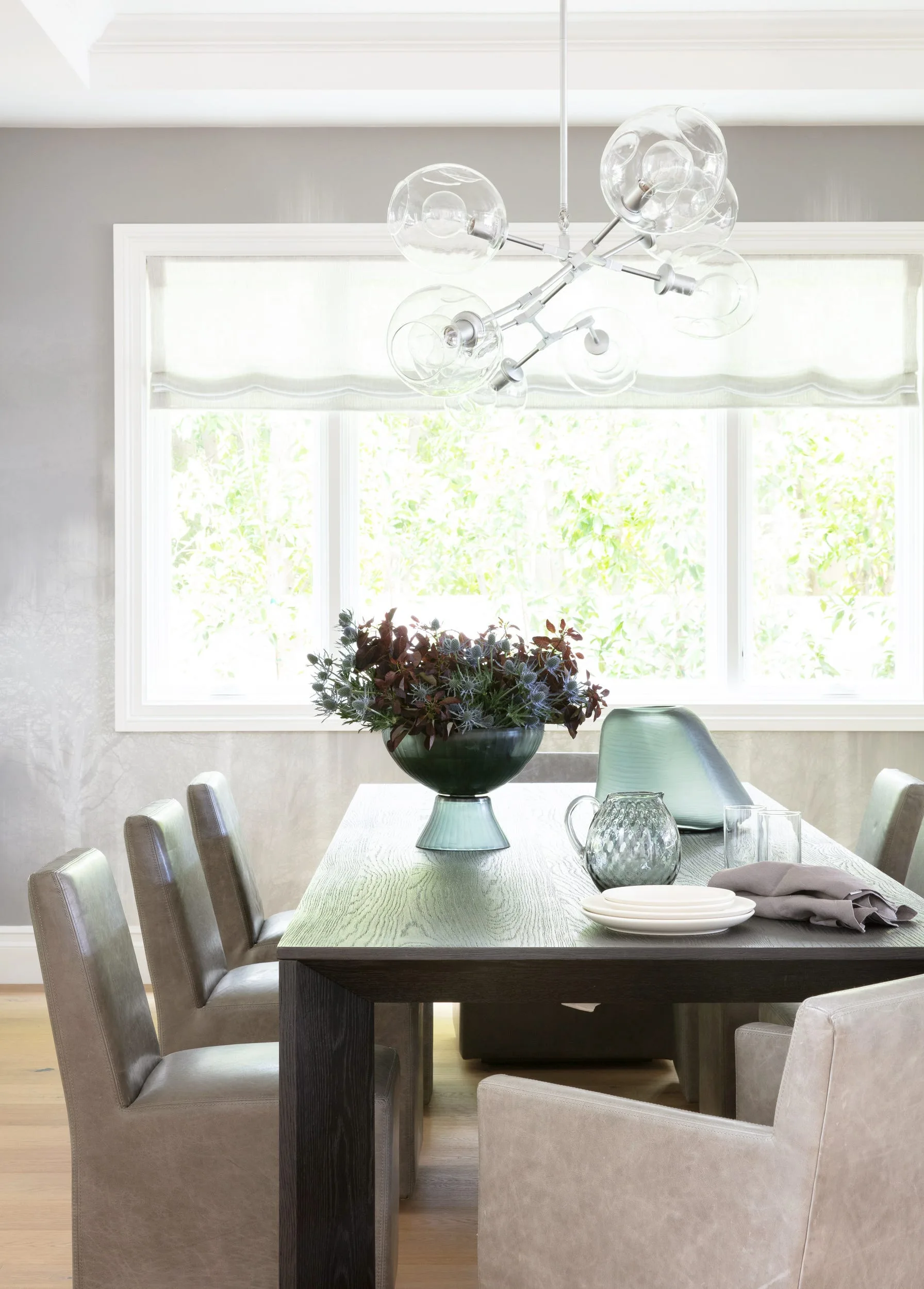 A dark wood dining table sits beneath a modern glass globe chandelier in a bright grey room with upholstered chairs.