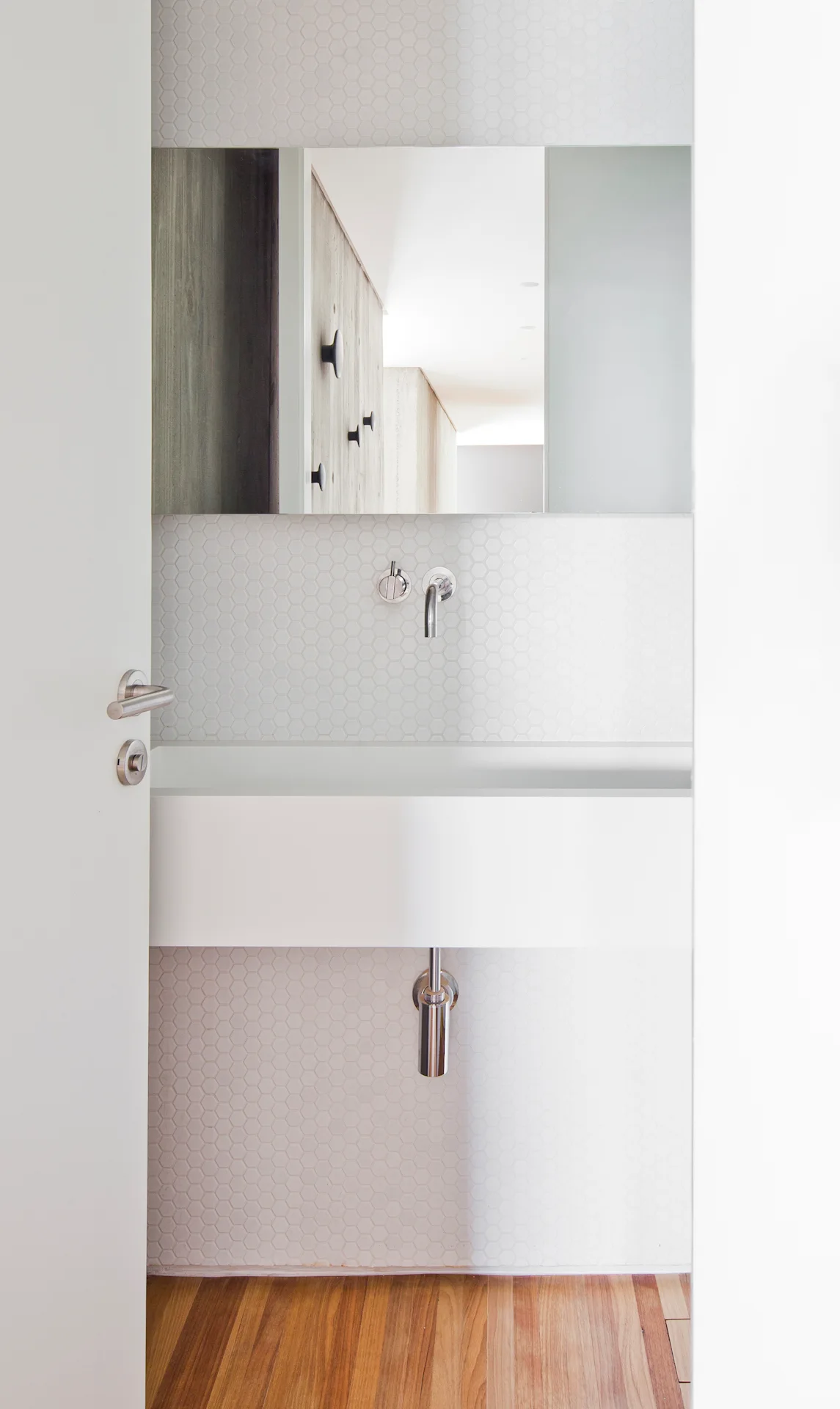 A white door opens to reveal a minimalist powder room with a white wall-mounted basin, chrome fixtures, and small hexagonal tile walls.