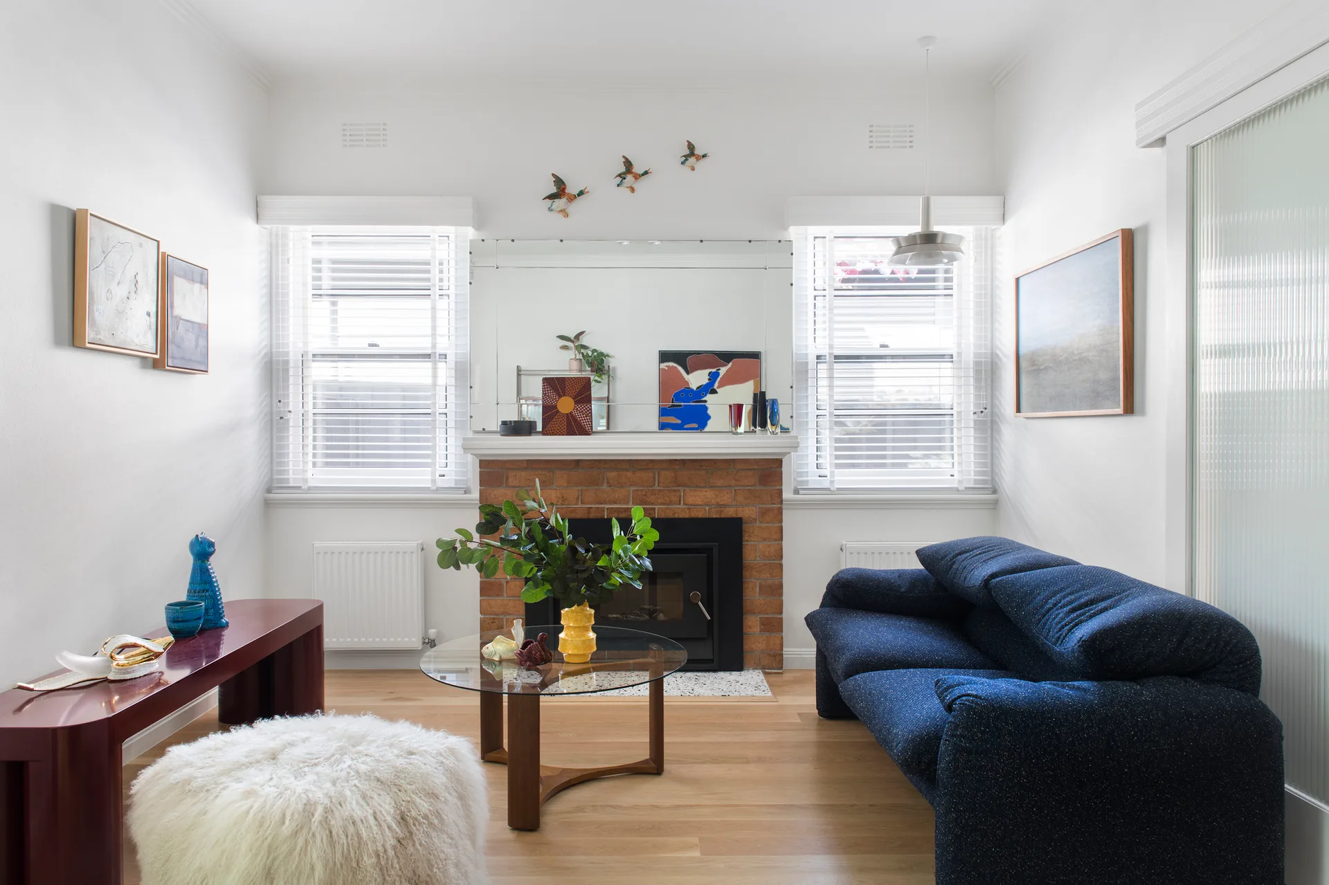 A bright living room with a central brick fireplace, a blue fabric sofa, two windows with white blinds, and a round glass coffee table.